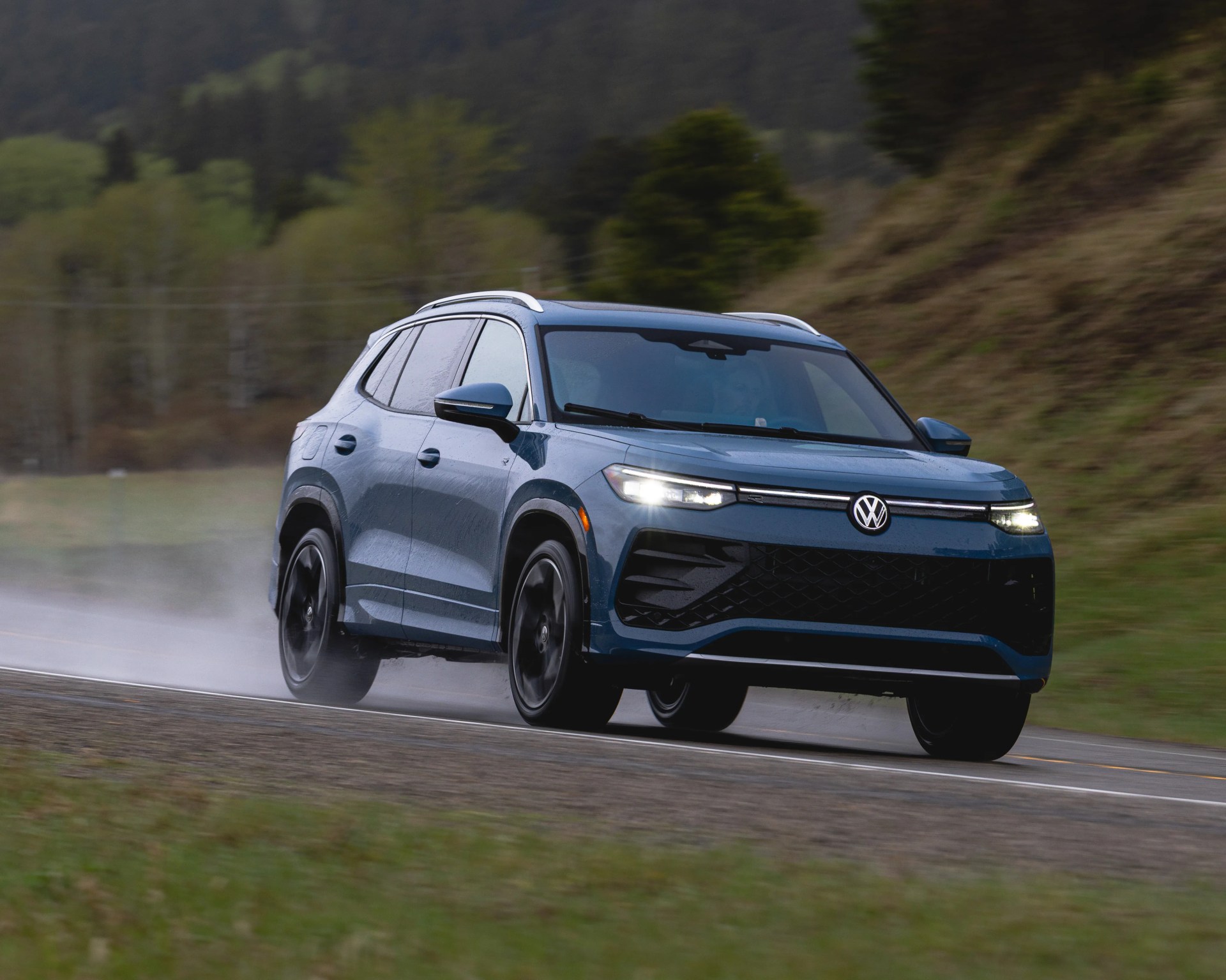 Blue Volkswagen SUV driving on a wet road with headlights on and forested hills in the background.