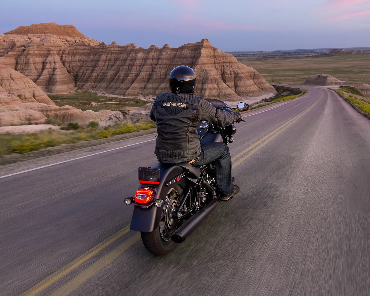 Person wearing a black Harley-Davidson jacket and helmet riding a black motorcycle on a winding road near rocky hills.