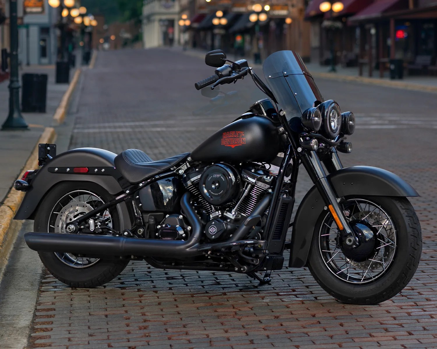 Matte black Harley-Davidson motorcycle with windshield and dual front headlights parked on a cobblestone street.
