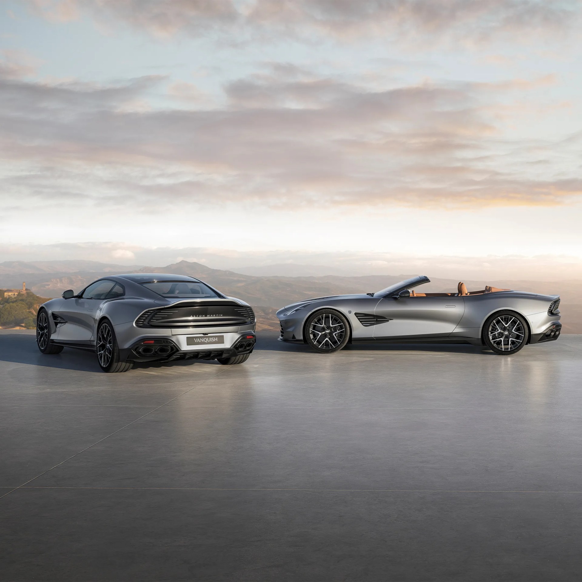 Two silver Aston Martin Vanquish cars, one coupe and one convertible, parked side by side on a concrete surface with mountains and a cloudy sky in the background.