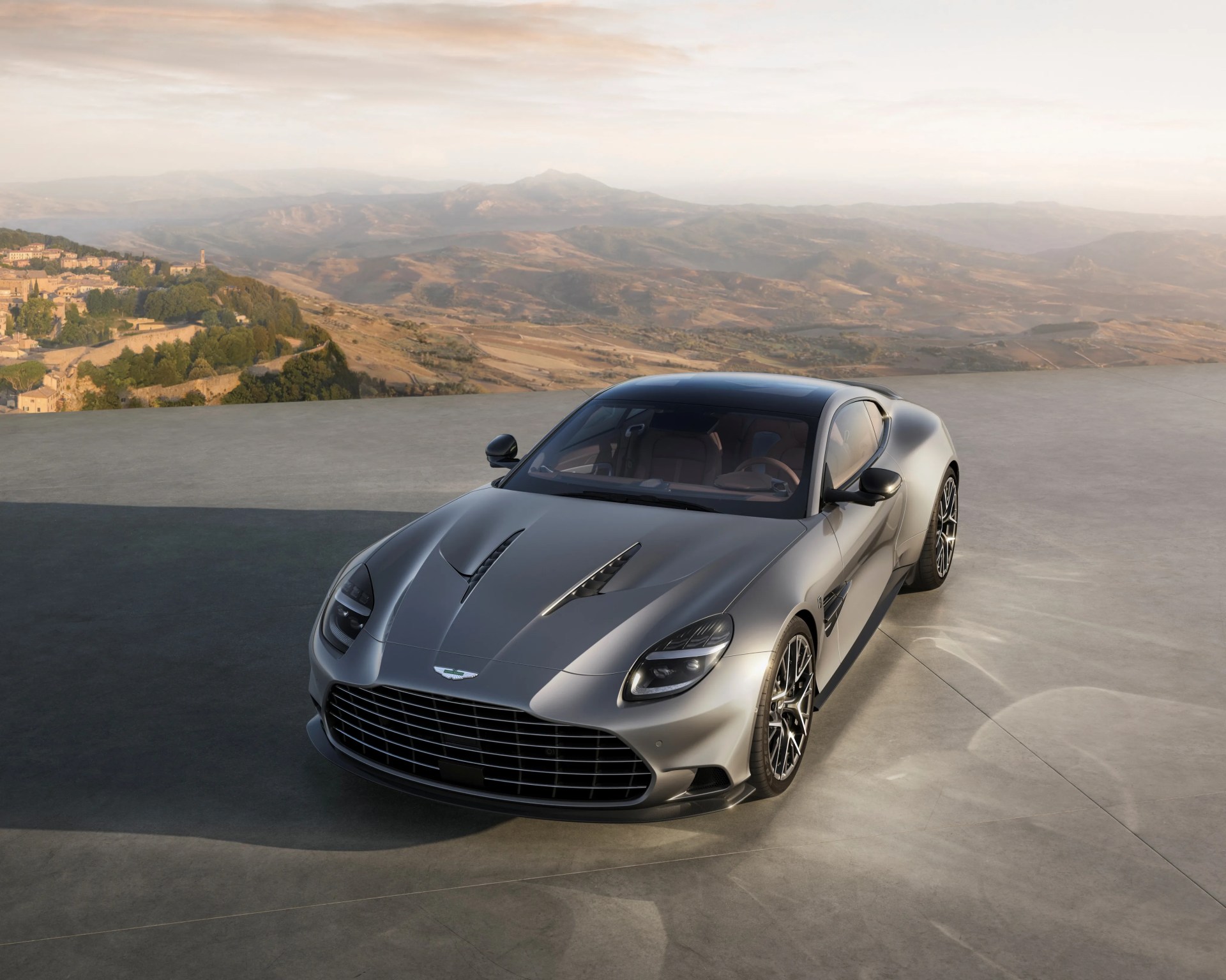 Silver Aston Martin sports car parked on a concrete surface with a mountainous landscape in the background.