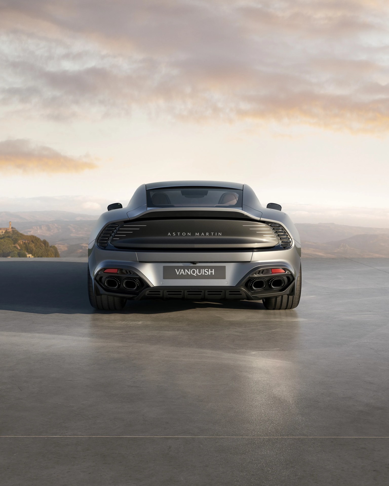 Rear view of a silver Aston Martin Vanquish with quad exhausts on a concrete surface under a cloudy sky.