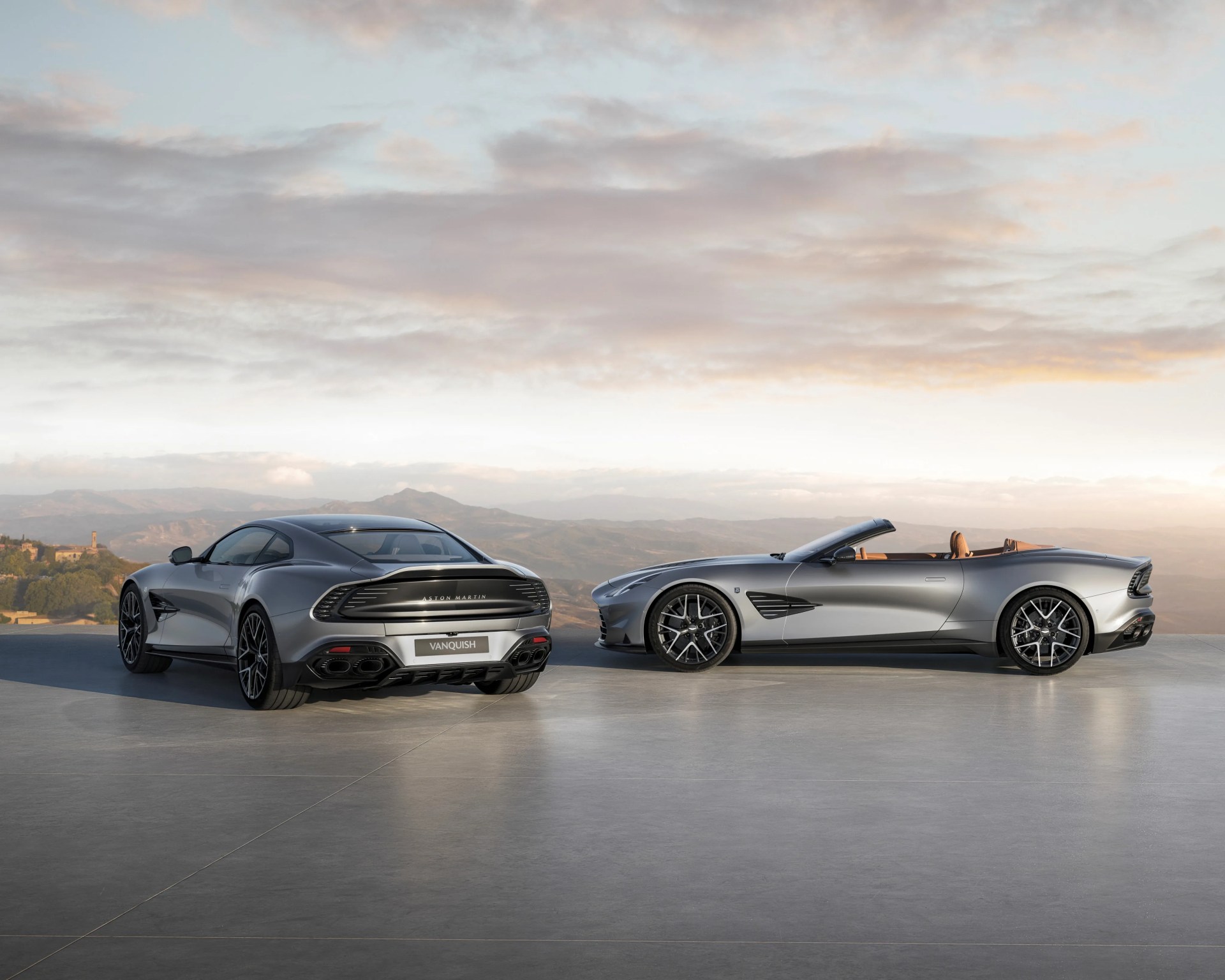 Two silver Aston Martin Vanquish cars, one coupe and one convertible, parked side by side on a concrete surface with mountains in the background.