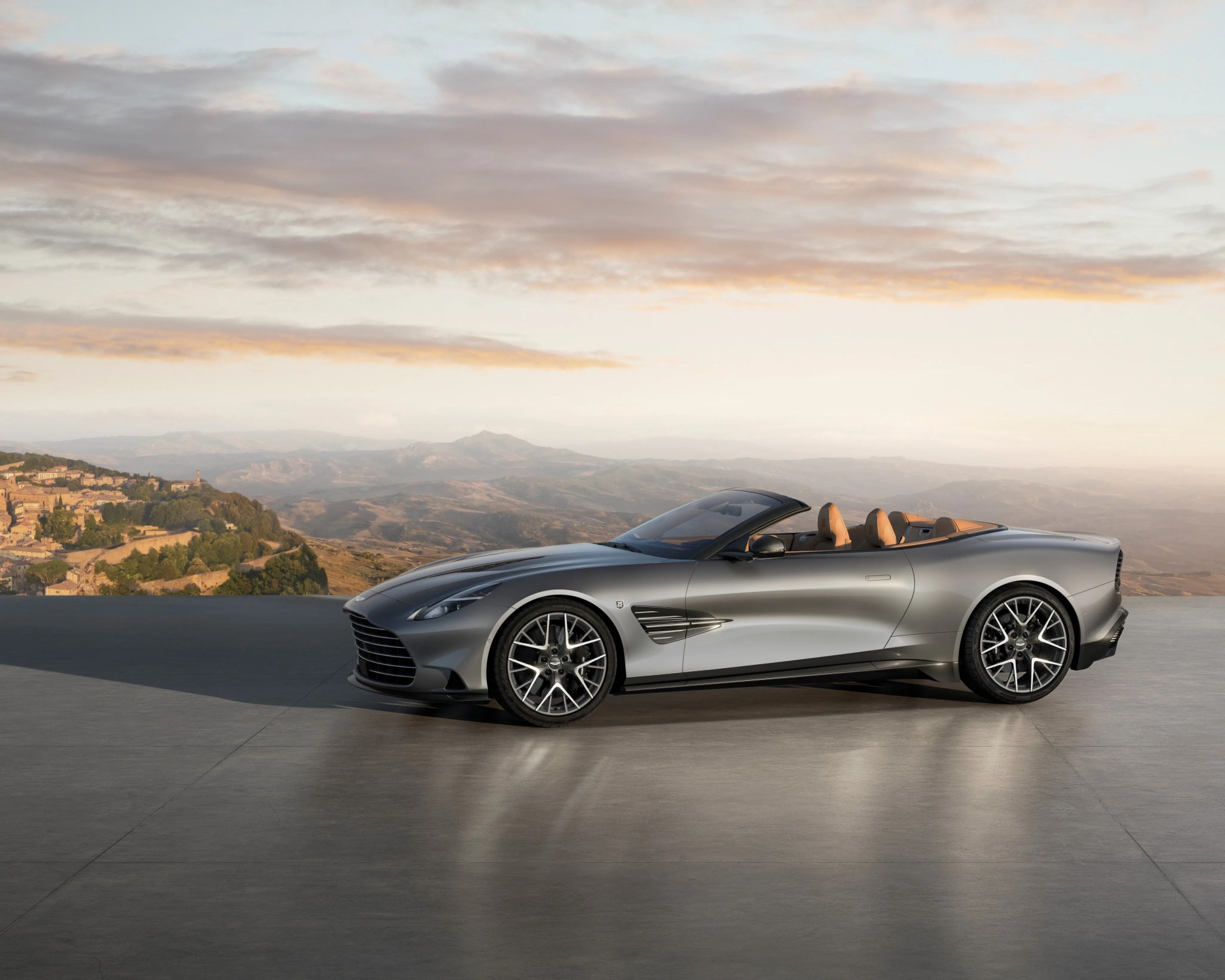 Silver convertible sports car with tan leather interior parked on a concrete surface with mountains and a village in the background.