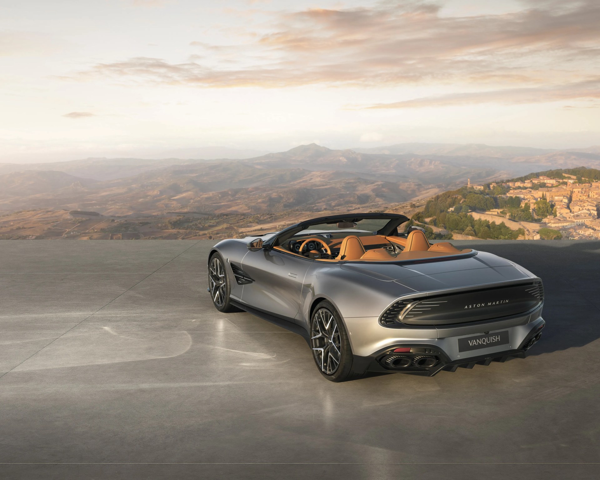Silver Aston Martin Vanquish convertible with tan leather interior parked on a concrete surface overlooking a mountainous landscape.