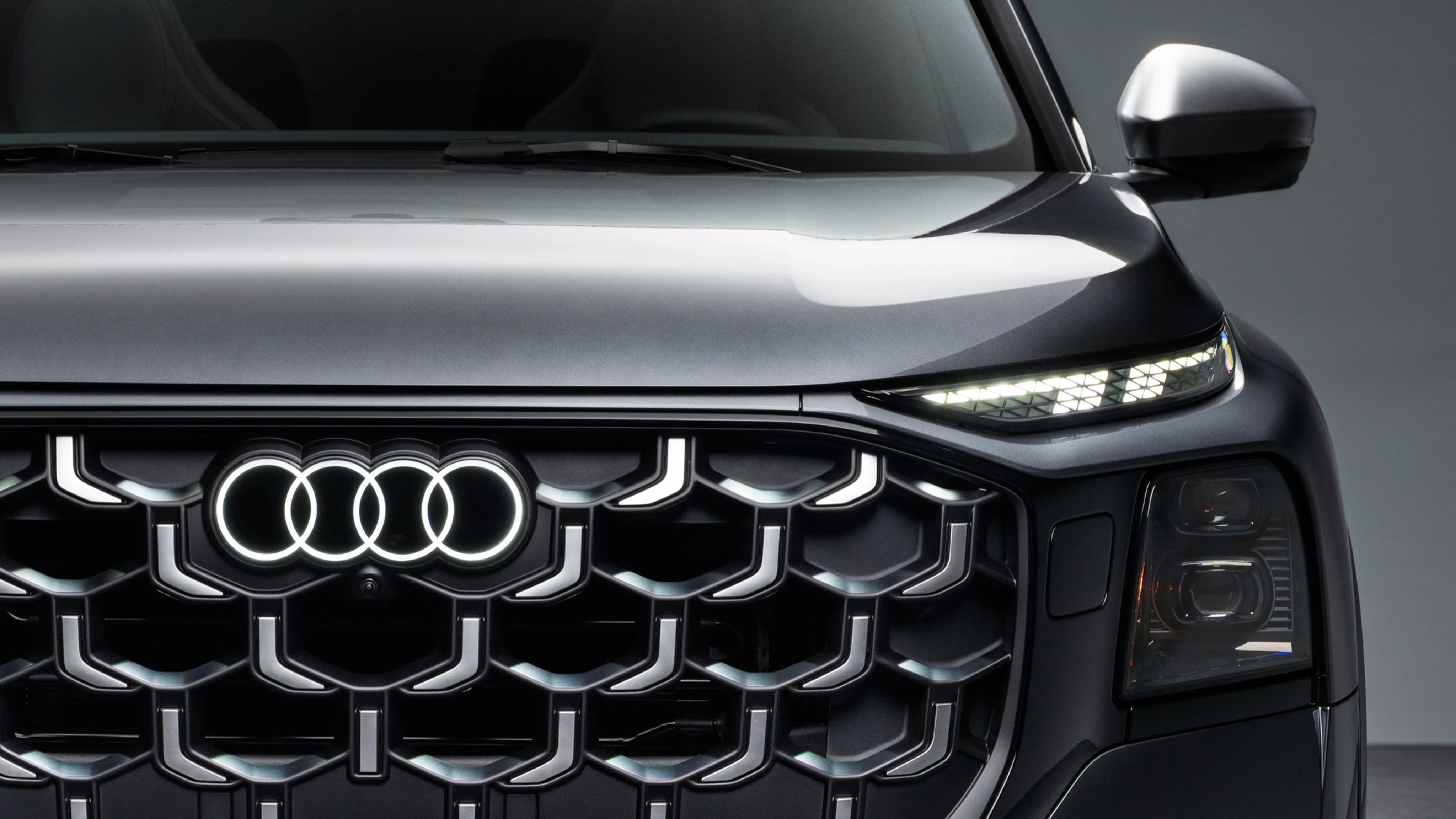 Front view of a dark gray Audi car showing illuminated logo, grille, and LED headlights.
