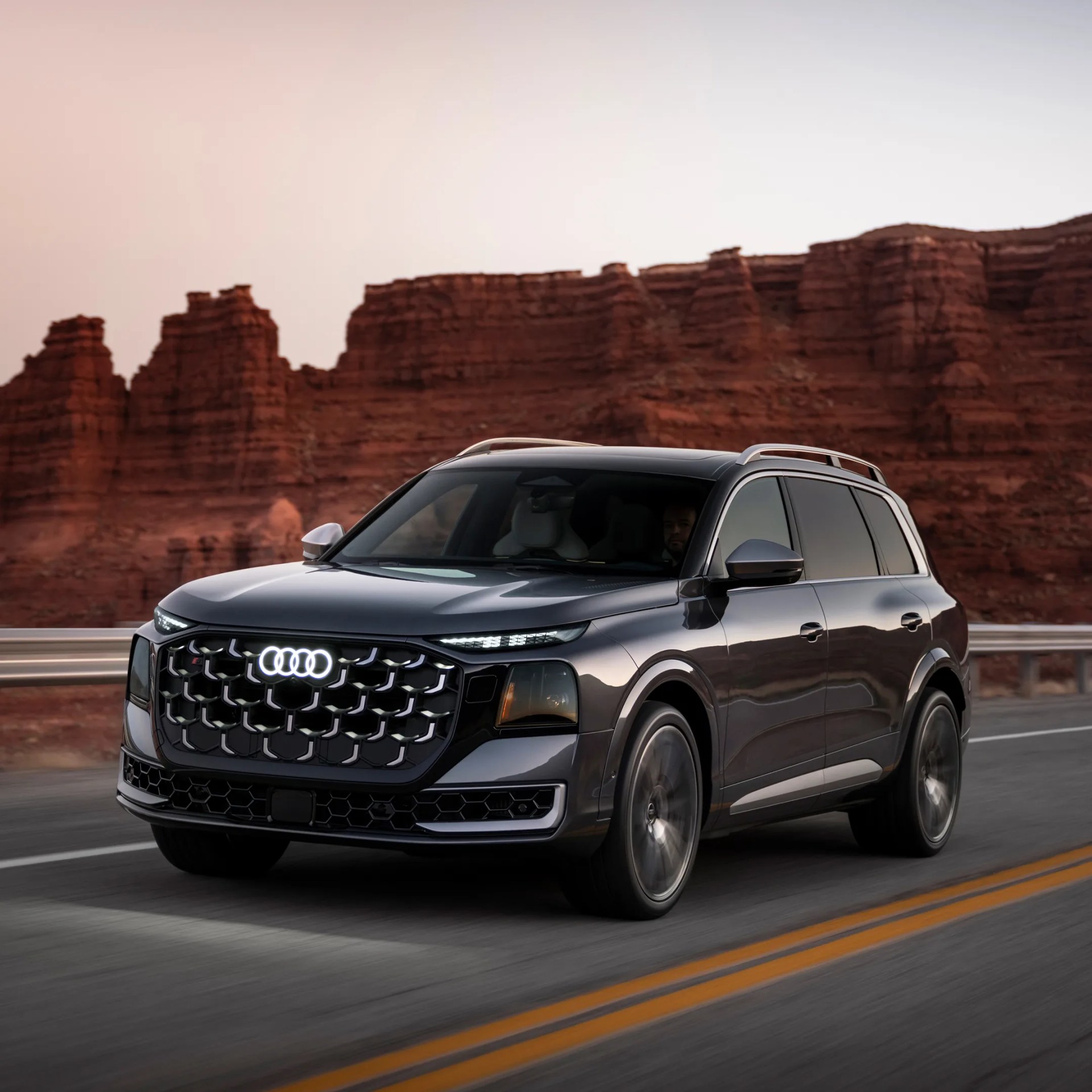 Black Audi SUV driving on a road with red rock cliffs in the background at sunset.