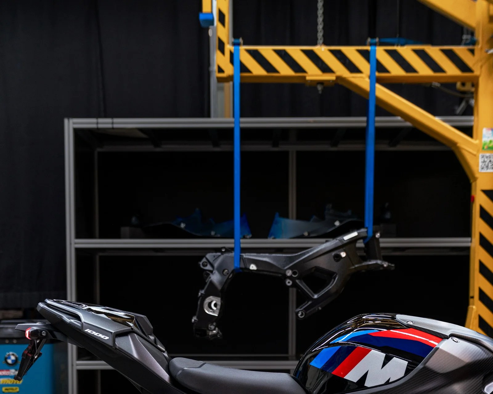 Rear section of a black motorcycle with "1000" decal and a blue, red, and white fuel tank, with a black frame suspended by blue straps.