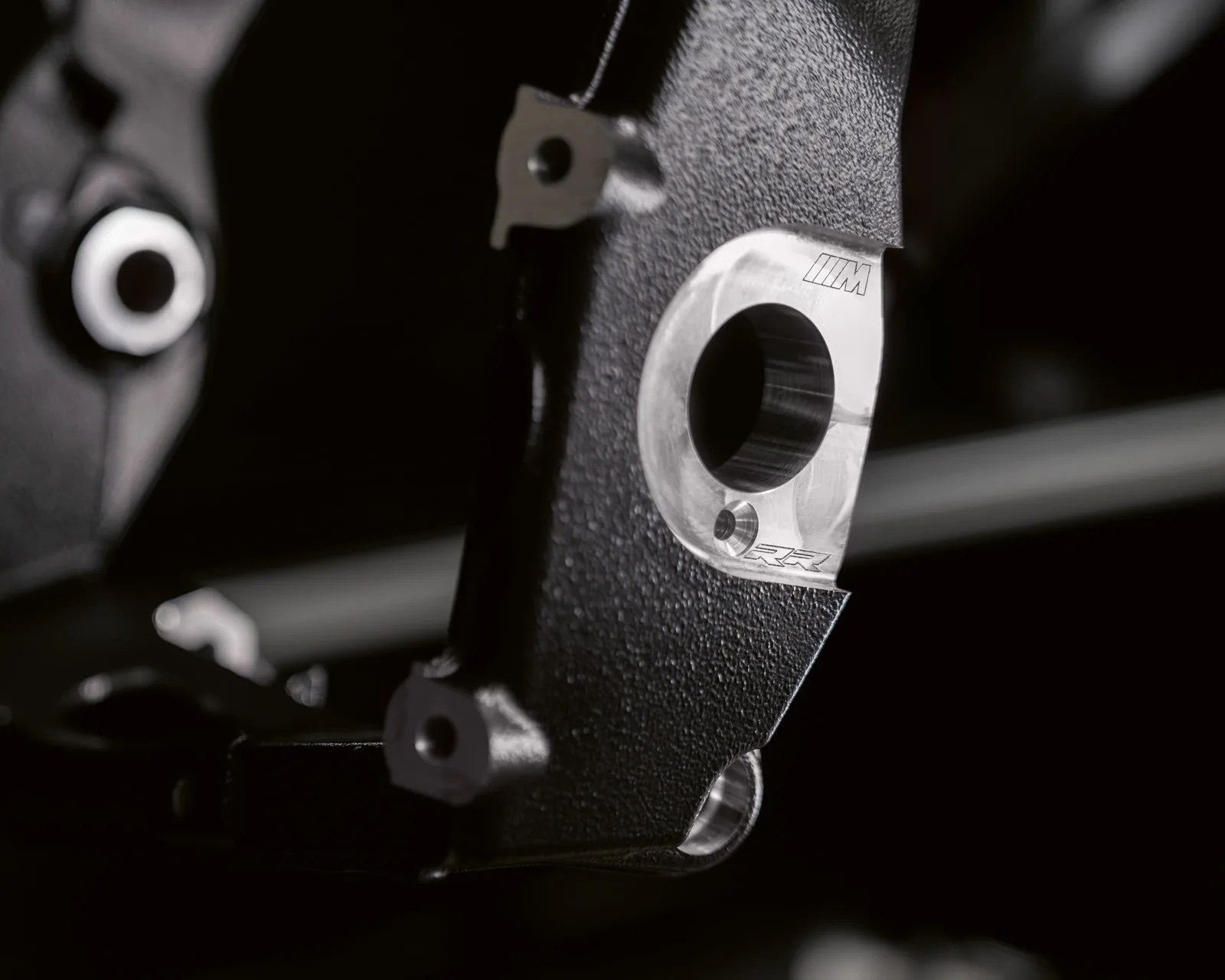 Close-up of a black textured metal component with a silver circular opening featuring engraved "M" and "RR" logos.