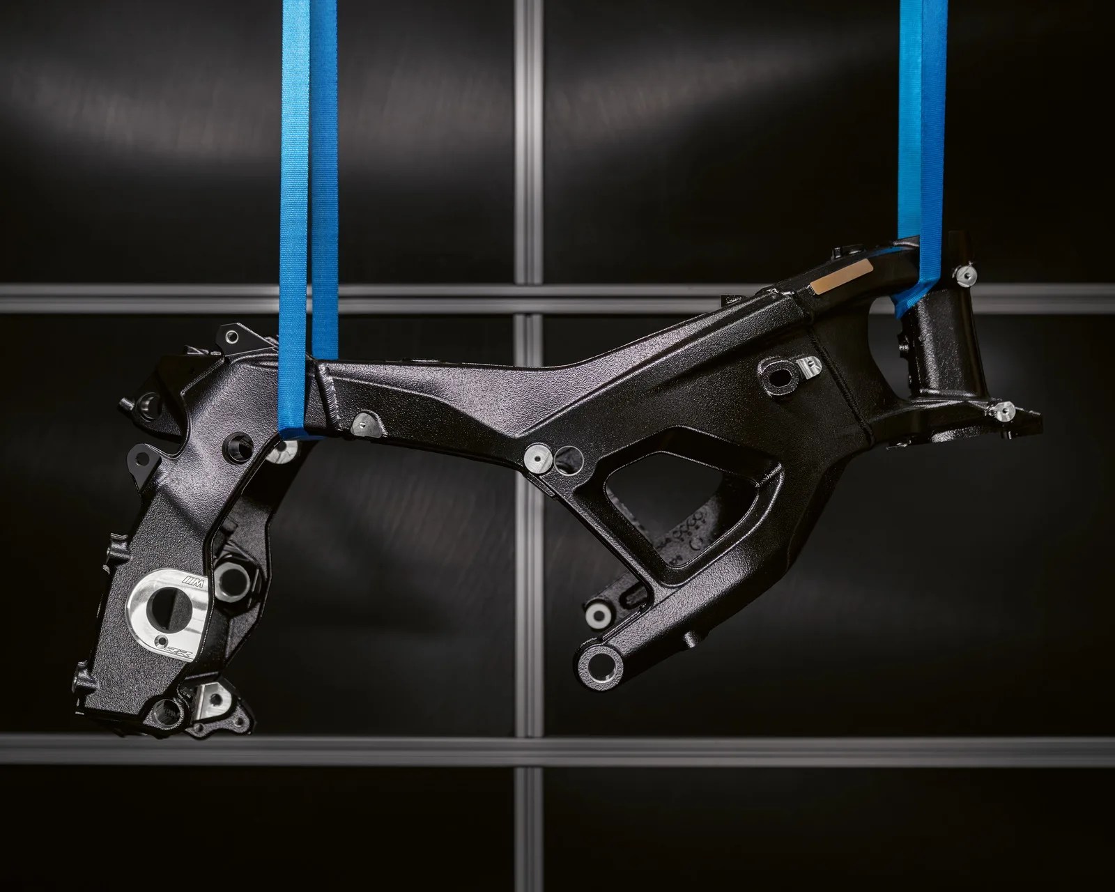 Black metal motorcycle frame suspended by blue straps against a dark background.
