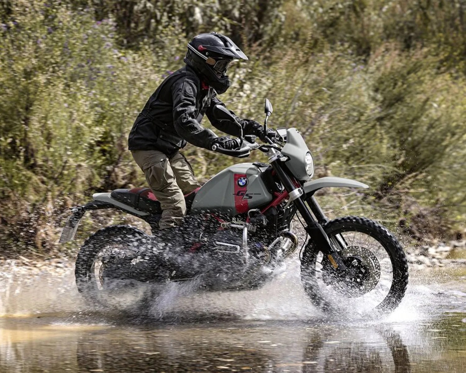 Person riding a gray and red BMW GS R1250 motorcycle splashing through shallow water with greenery in the background.