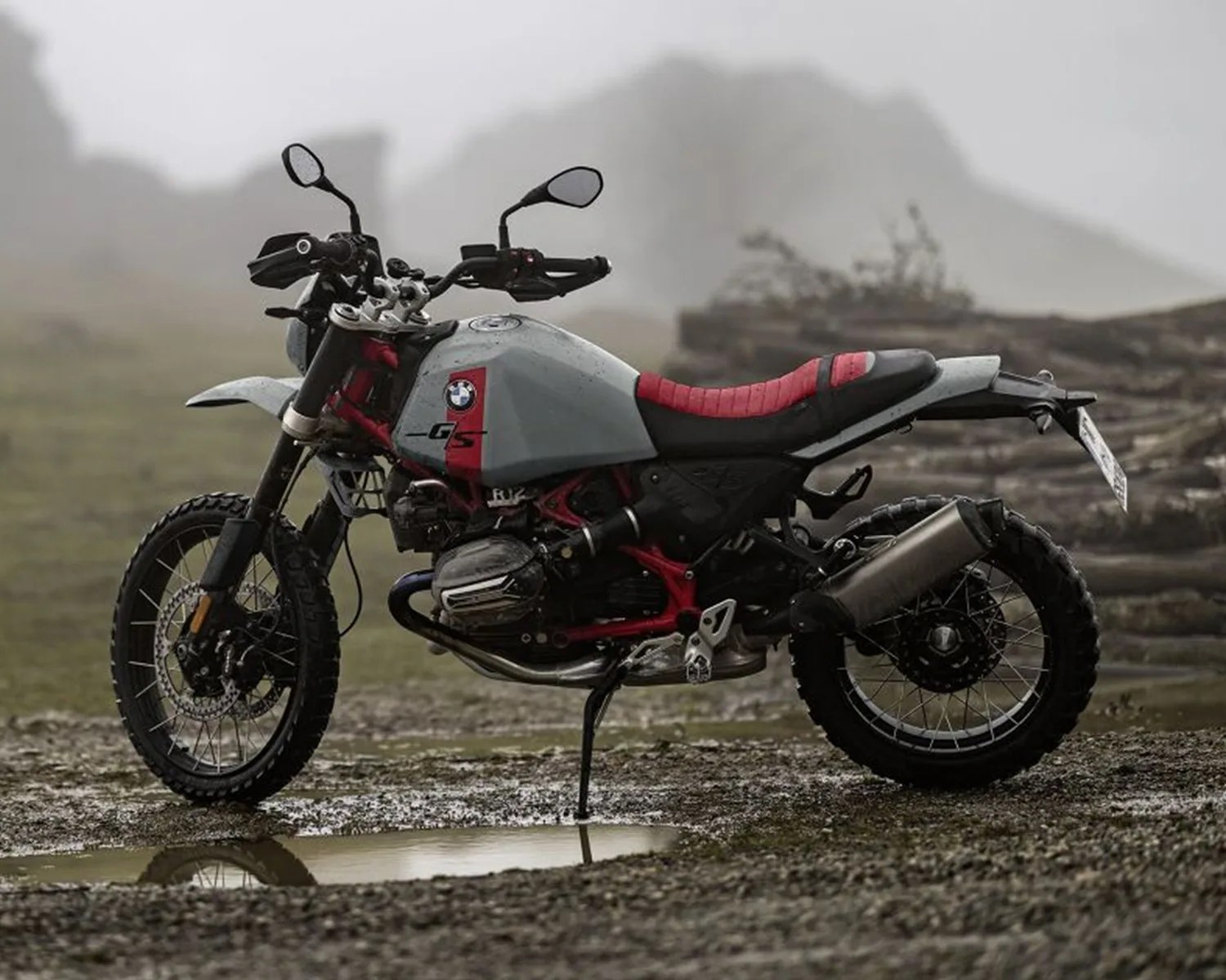 BMW GS motorcycle with gray and red body and knobby tires parked on muddy ground near a puddle.