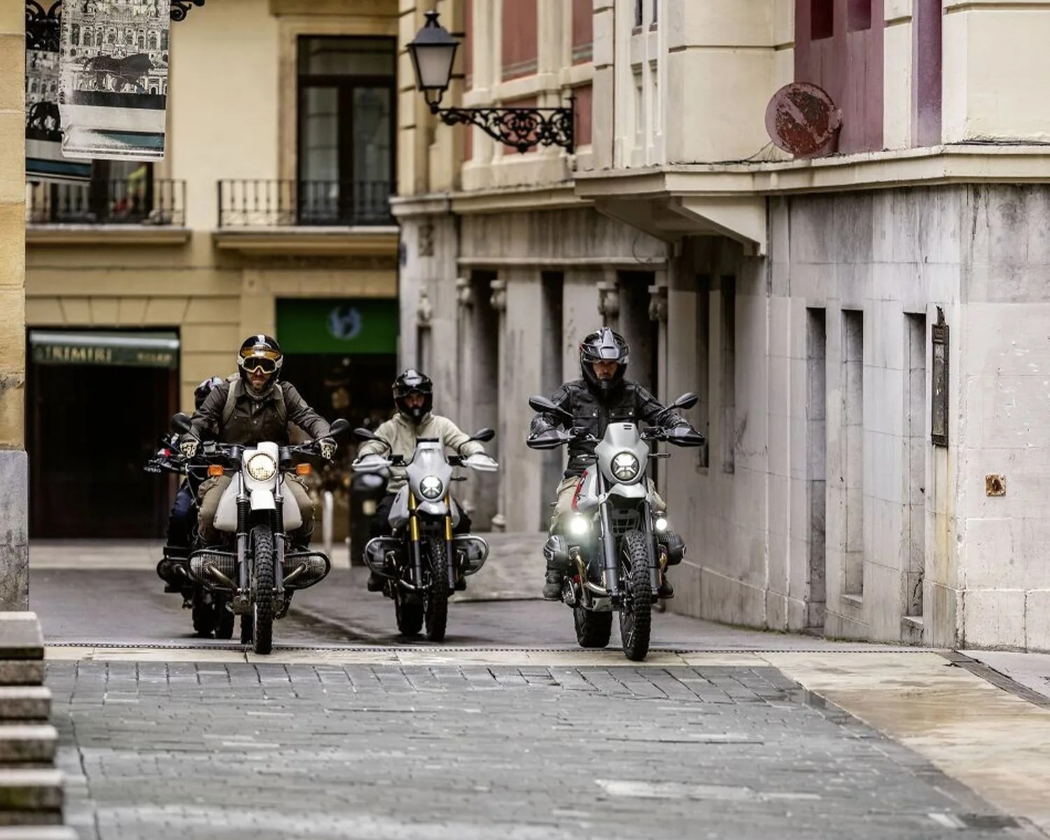 Three motorcyclists wearing helmets riding on a narrow cobblestone street between stone buildings.
