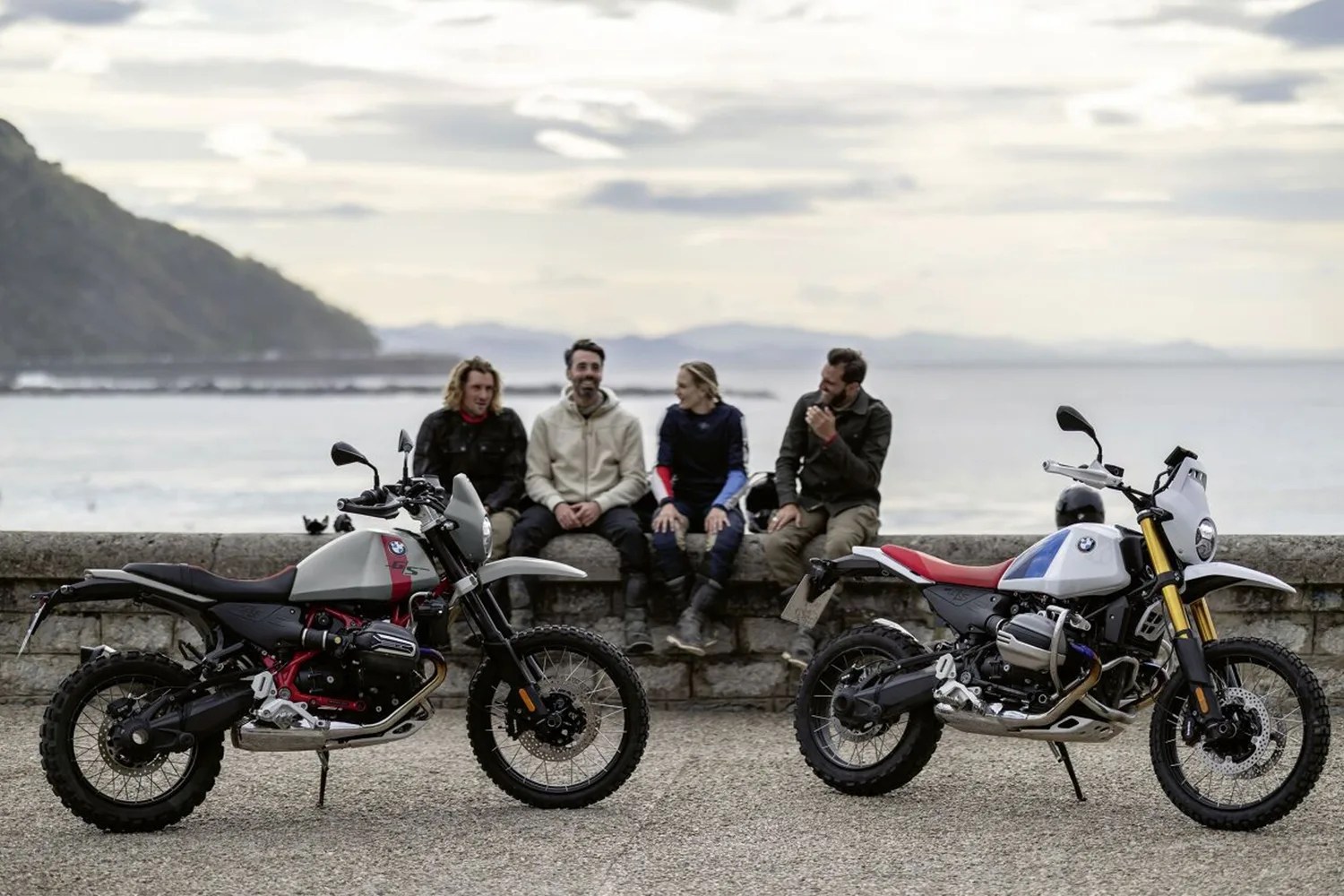 Two BMW GS motorcycles with knobby tires parked on gravel near a stone wall with four people sitting behind them by the water.