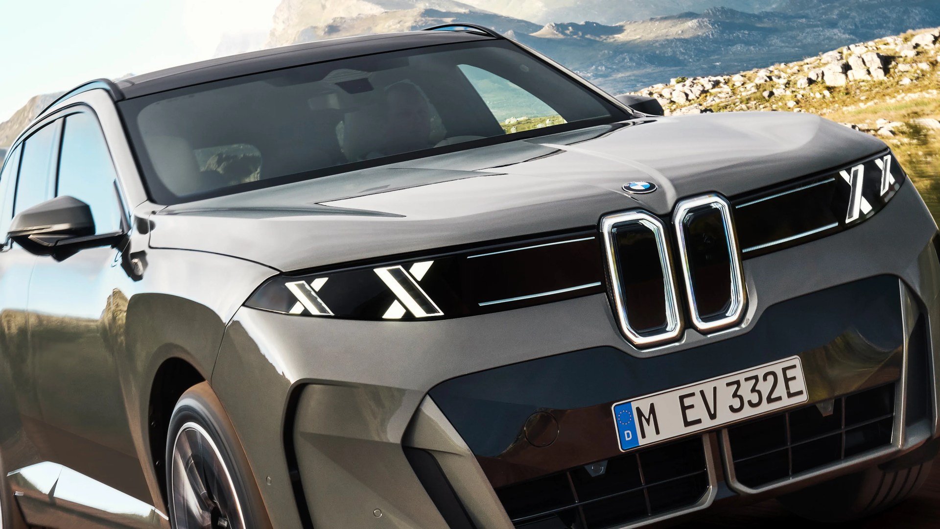 Front view of a gray BMW SUV with illuminated X-shaped LED headlights and a kidney grille, set against a mountainous background.