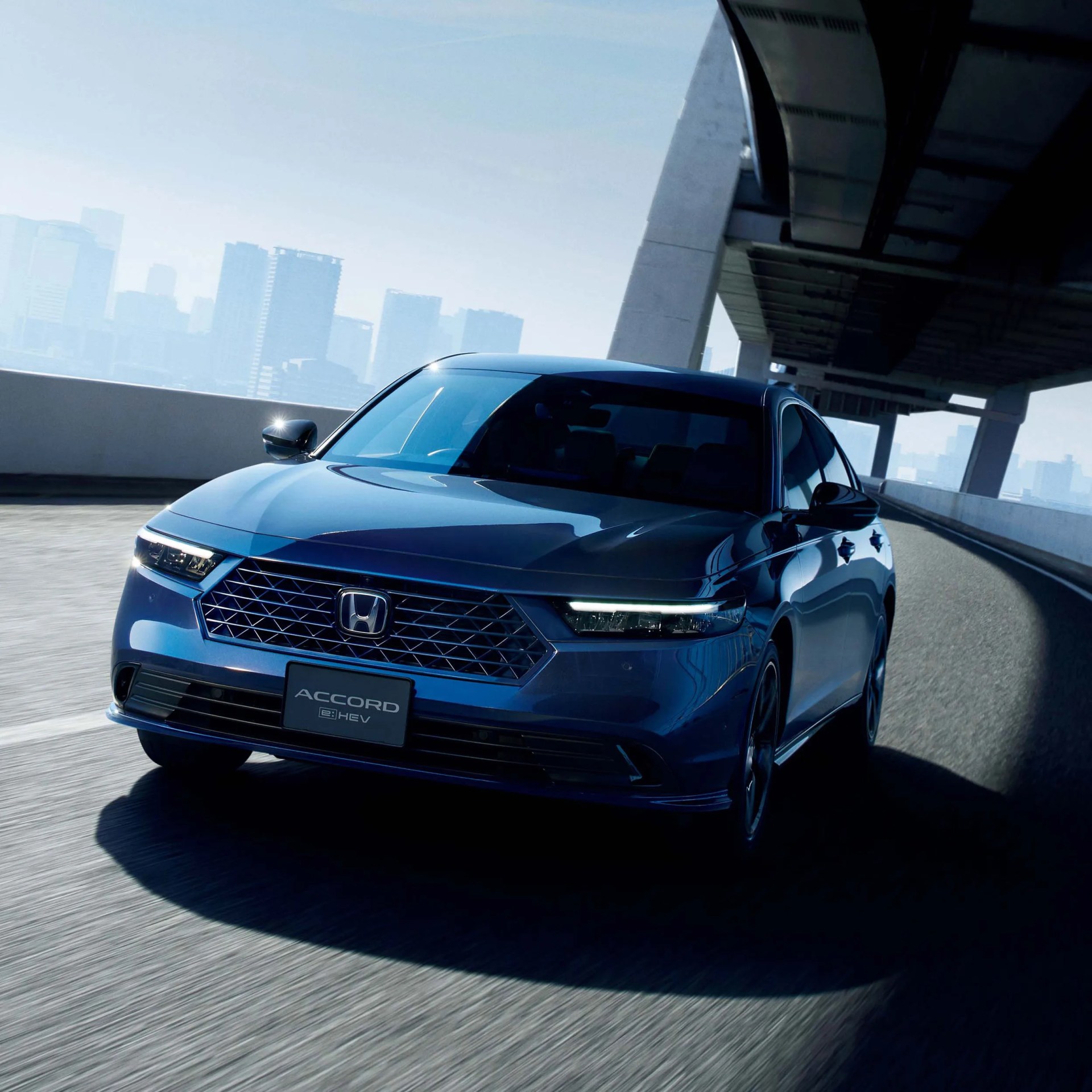 Blue Honda Accord e:HEV sedan driving on an elevated urban road with city skyline in the background.