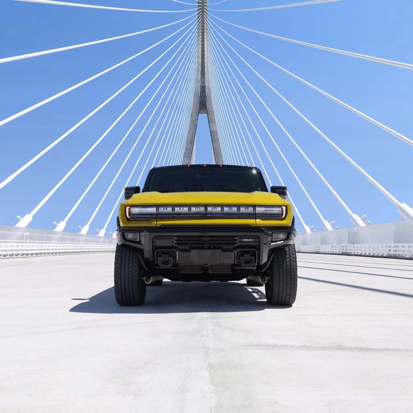 Yellow and black Hummer EV truck centered on a white cable-stayed bridge under a clear blue sky.