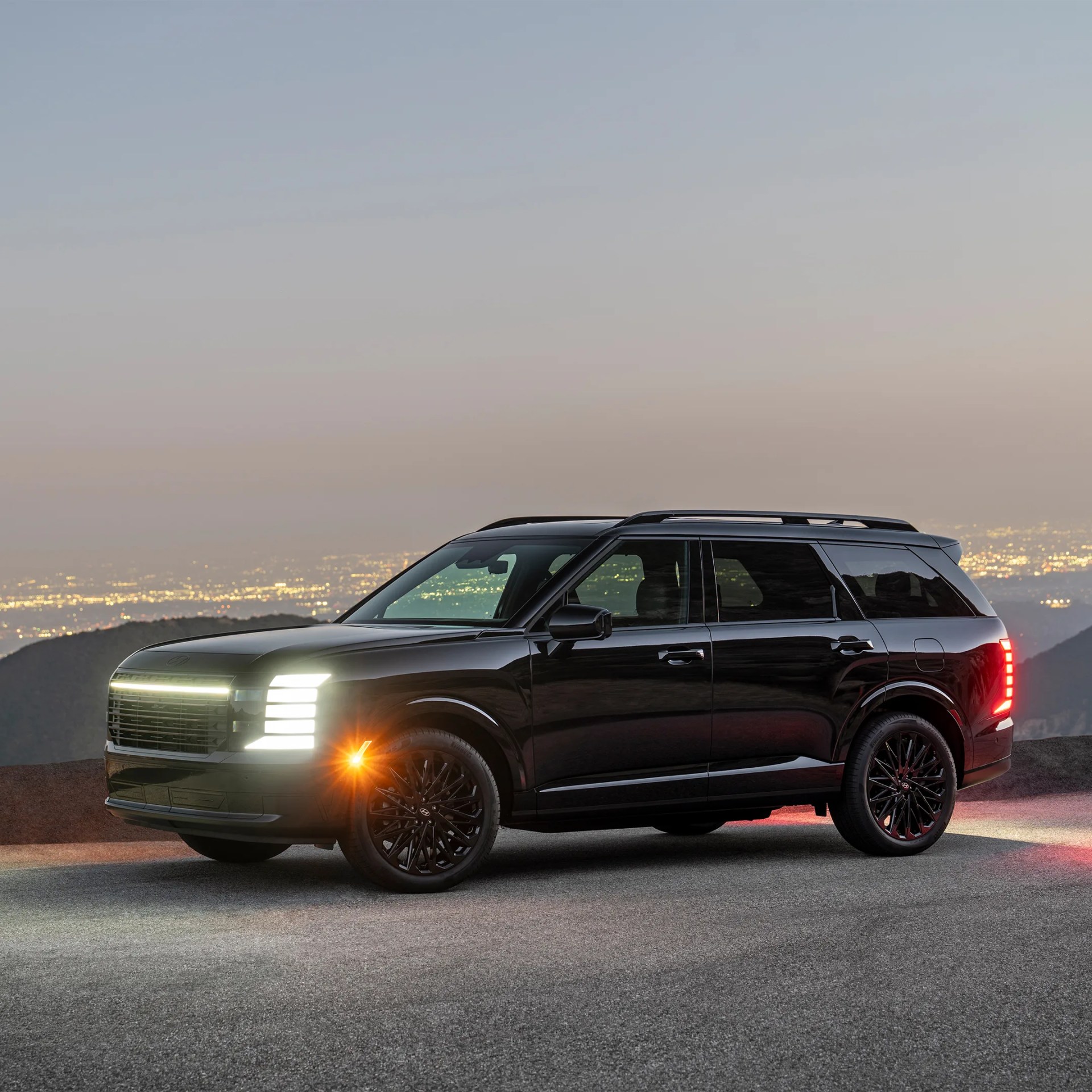 Black Hyundai SUV with illuminated headlights and taillights parked on a road at dusk with city lights in the background.