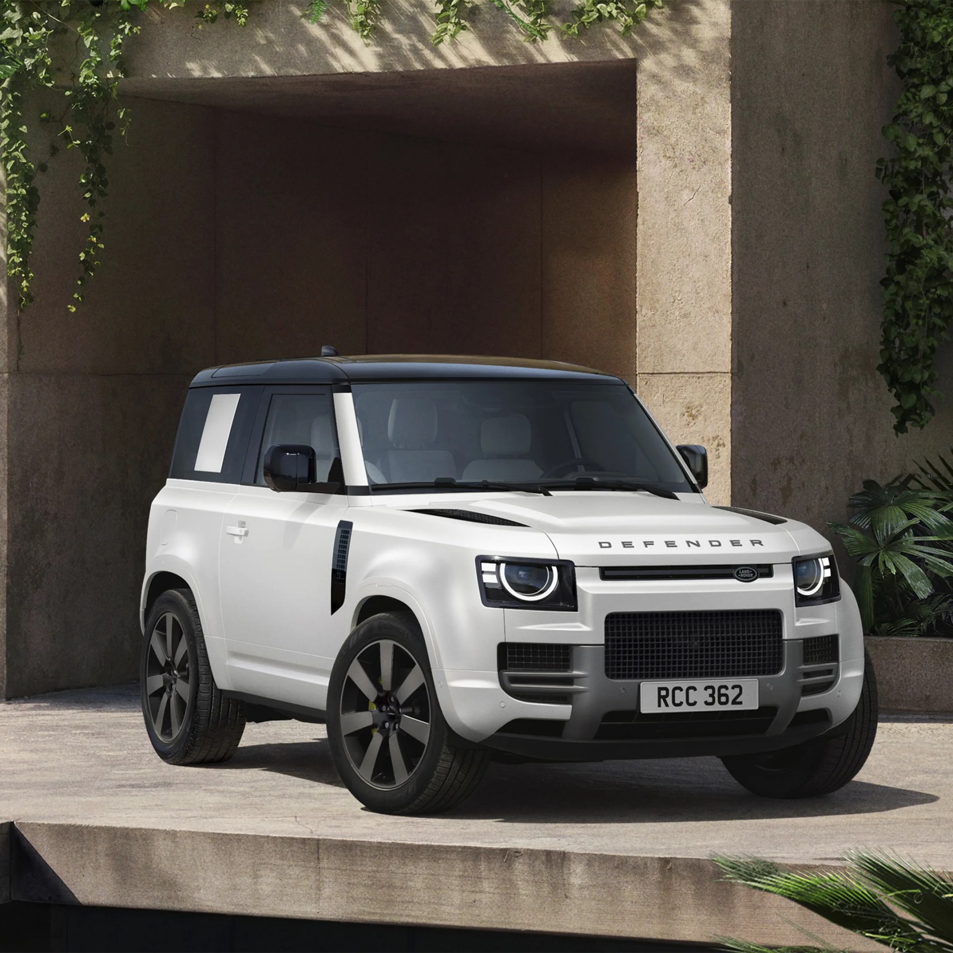 White Land Rover Defender with black roof and wheels parked on a concrete platform near a stone wall with greenery.