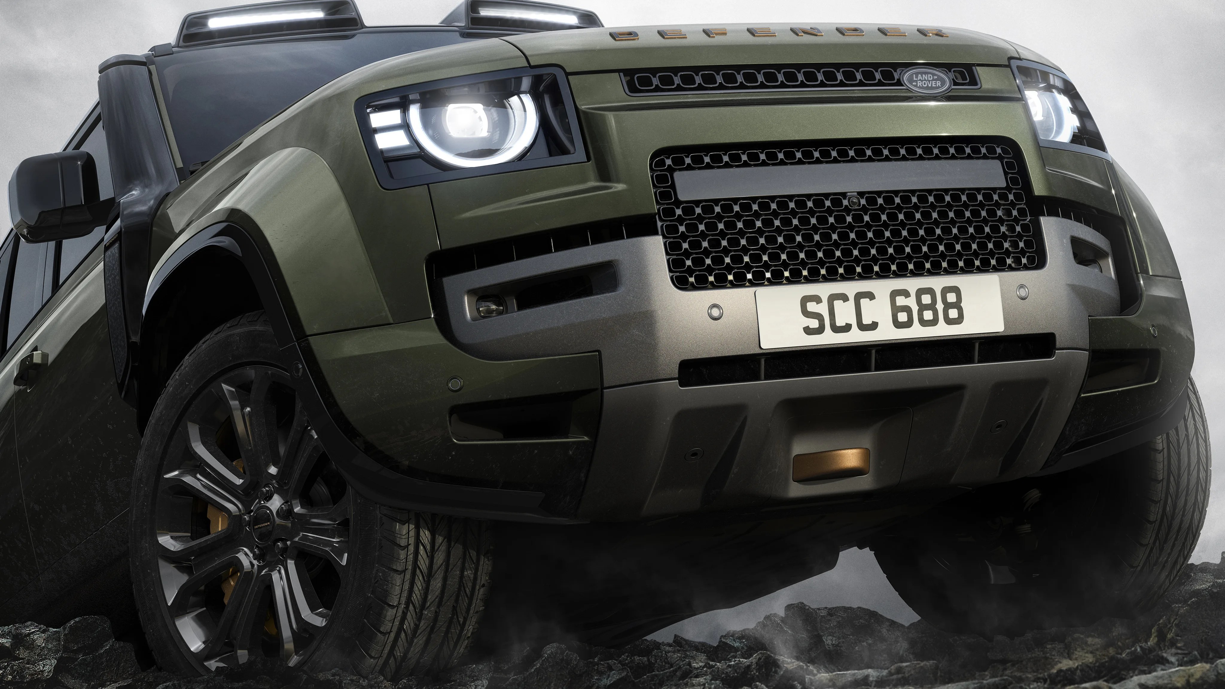 Front view of an olive green Land Rover Defender with illuminated headlights on rocky terrain.