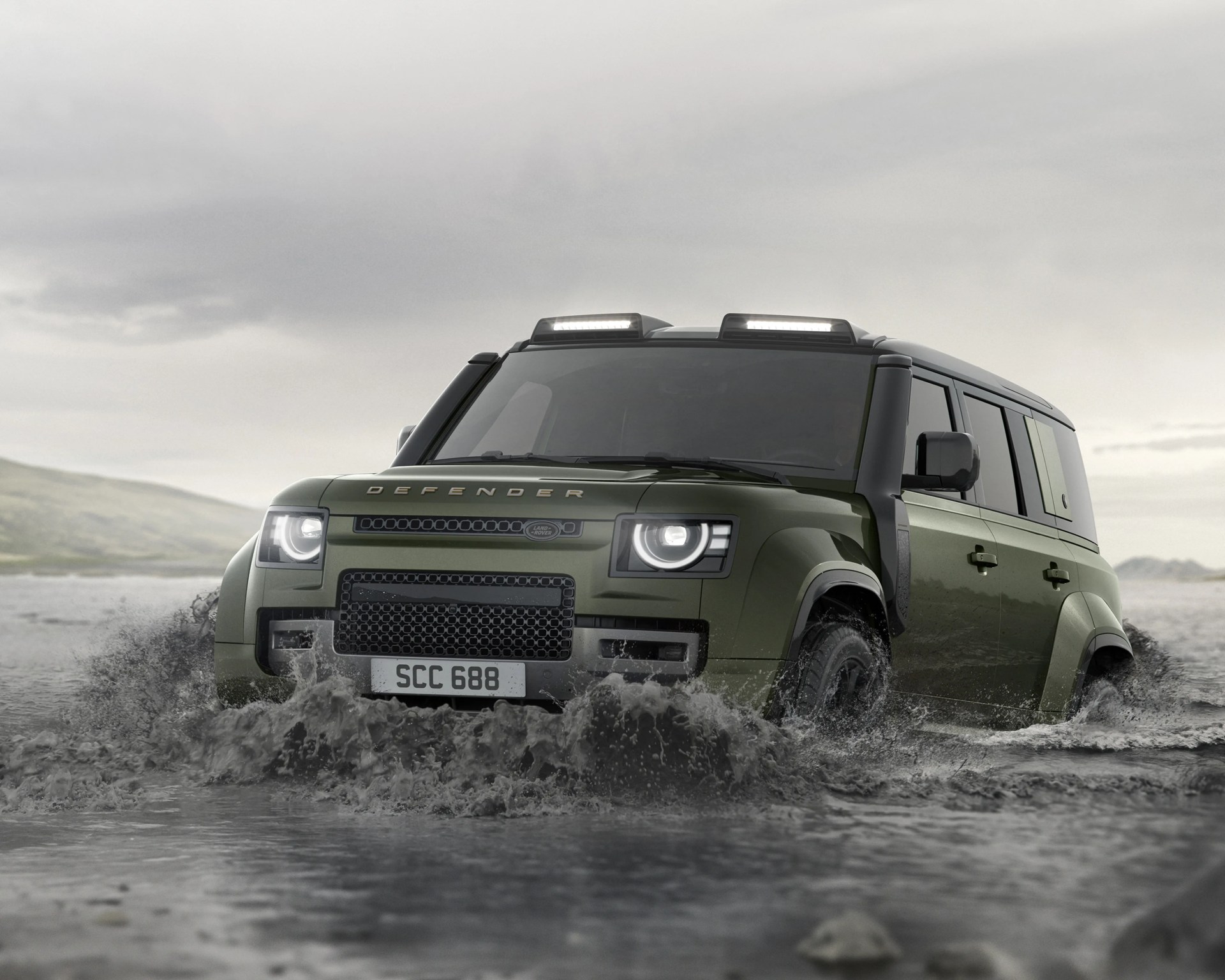 Green Land Rover Defender driving through water splashing around its front wheels under a cloudy sky.