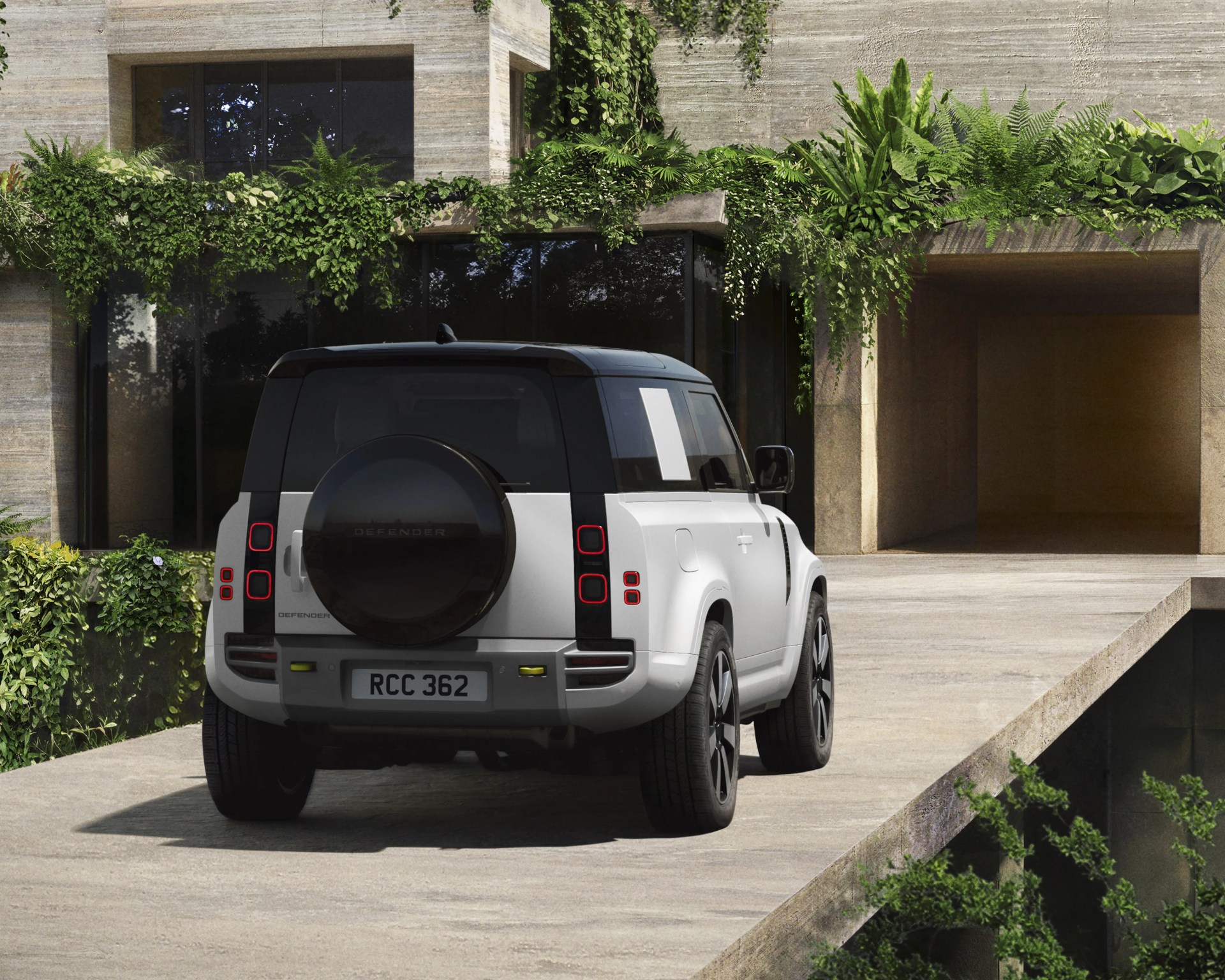 White Land Rover Defender SUV with black roof and spare tire mounted on the rear, parked on a concrete driveway with greenery above.