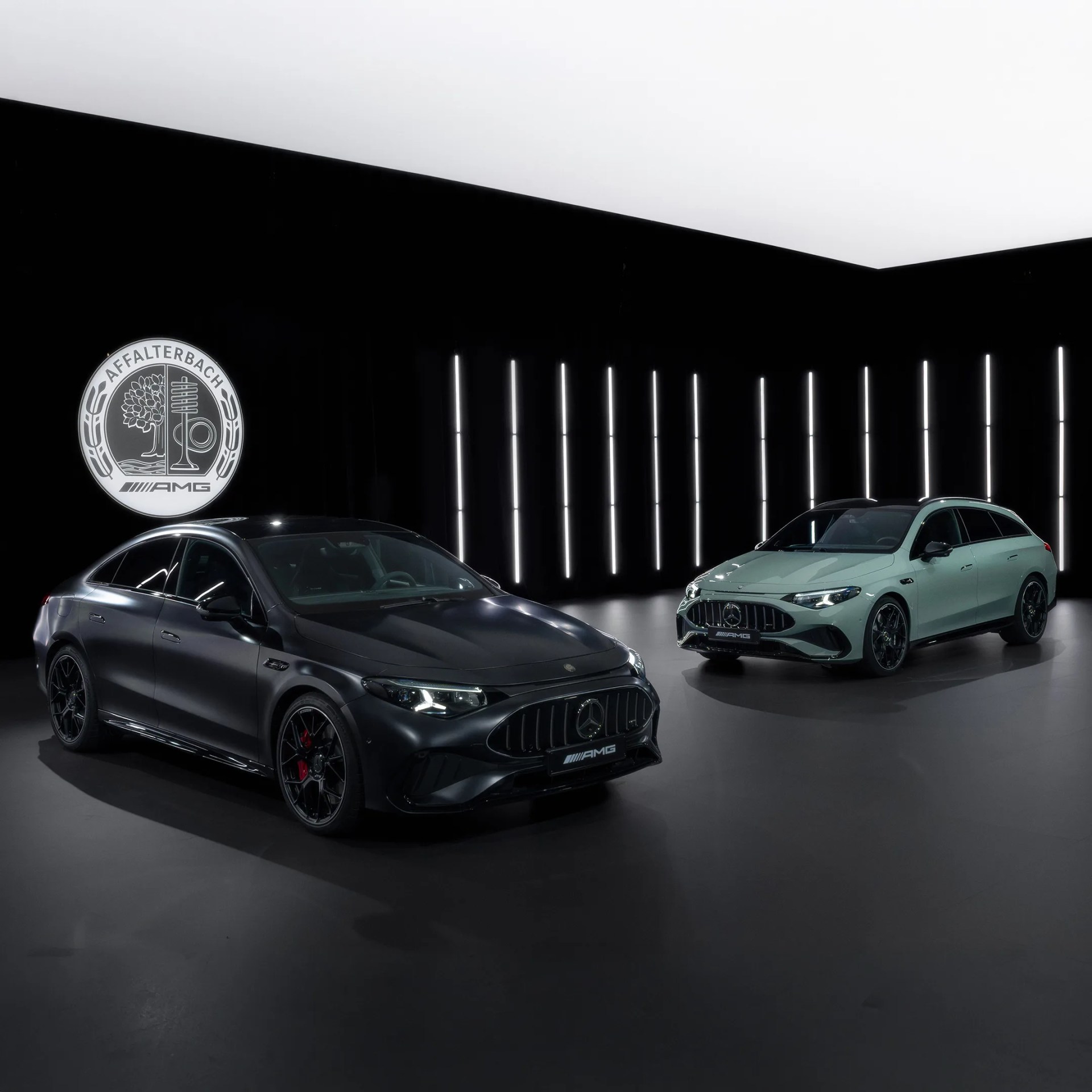Two Mercedes-AMG cars, one matte black sedan and one light green station wagon, displayed in a dark room with vertical white lights and an AMG logo on the wall.