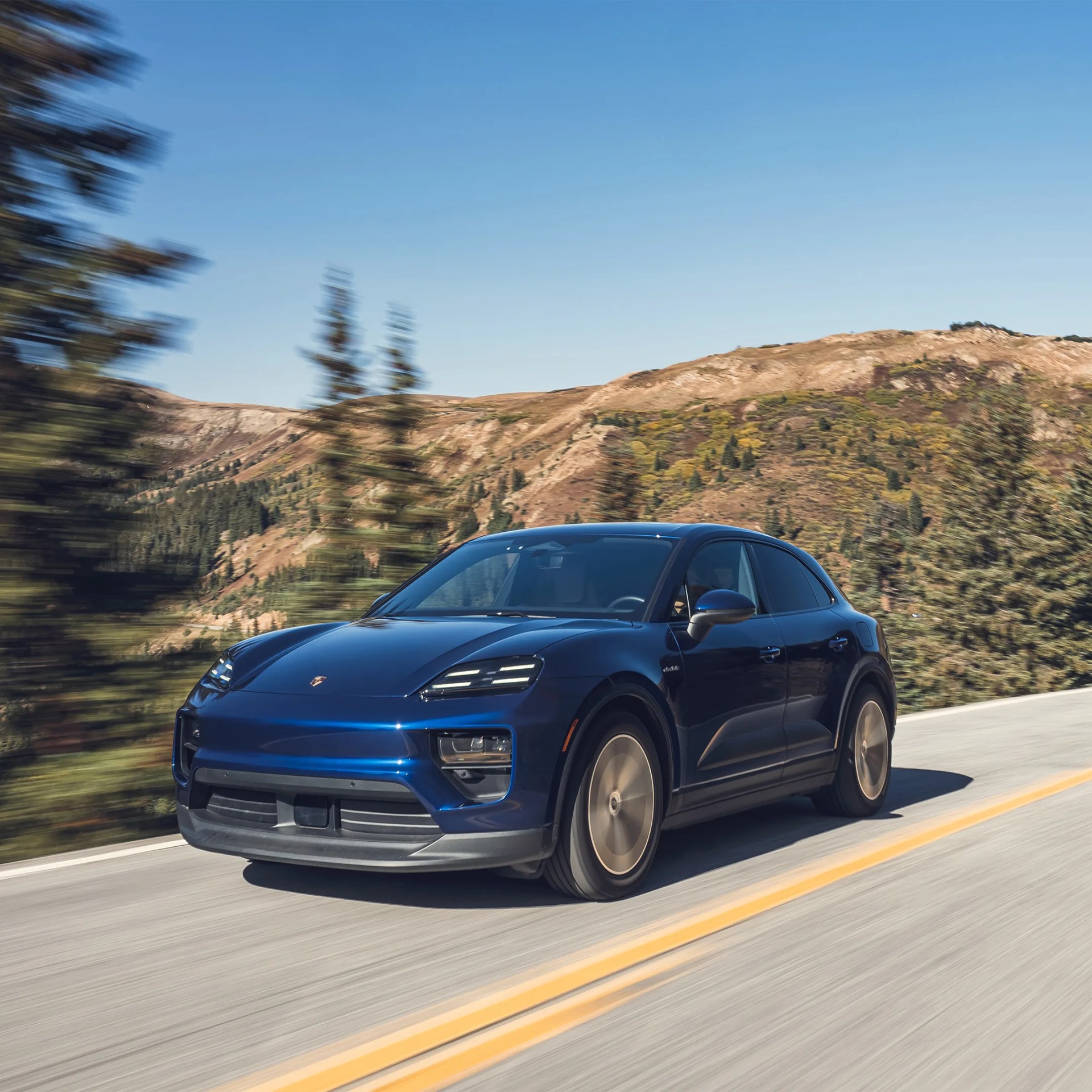 Blue Porsche electric SUV driving on a mountain road with blurred trees and hills in the background.