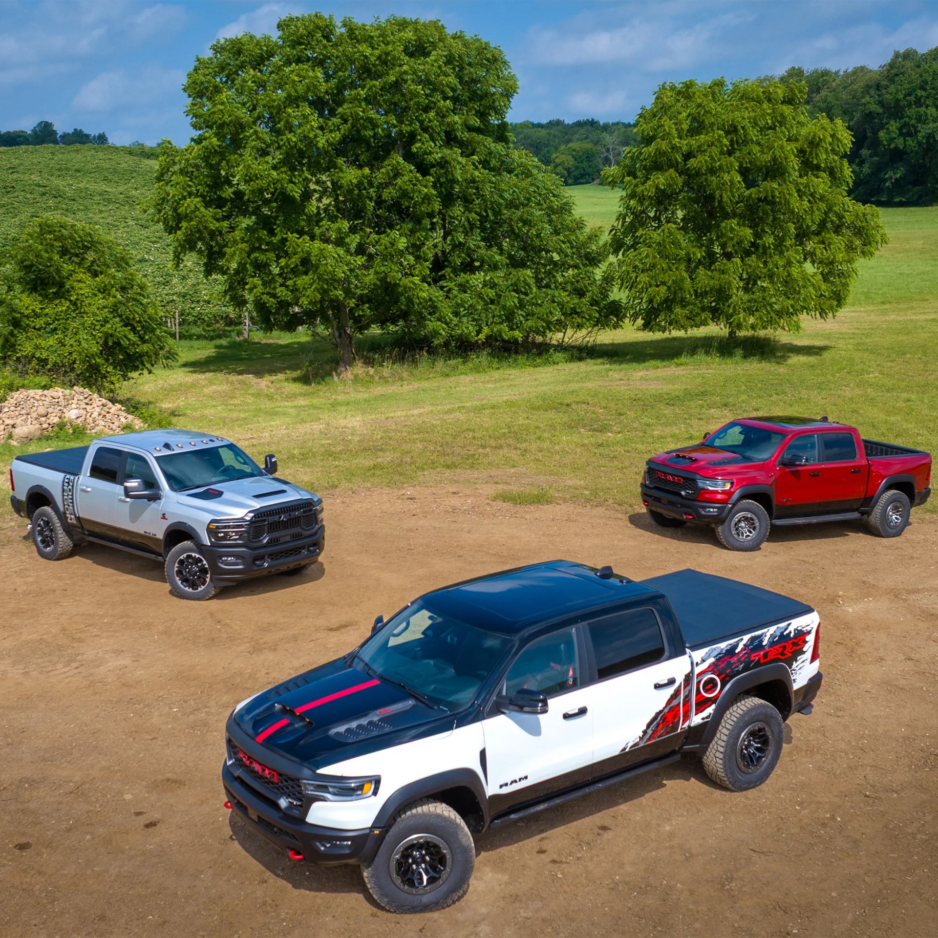 Three RAM pickup trucks in white, silver, and red parked on a dirt area with green trees and grass in the background.