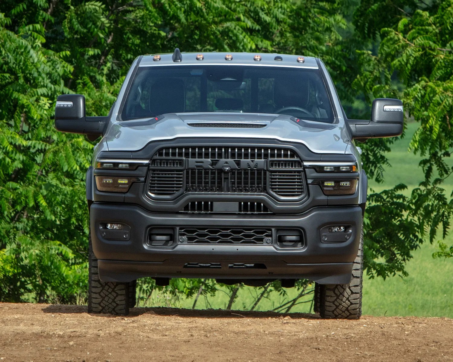 Front view of a silver RAM pickup truck with black grille and off-road tires on a dirt surface with green trees in the background.