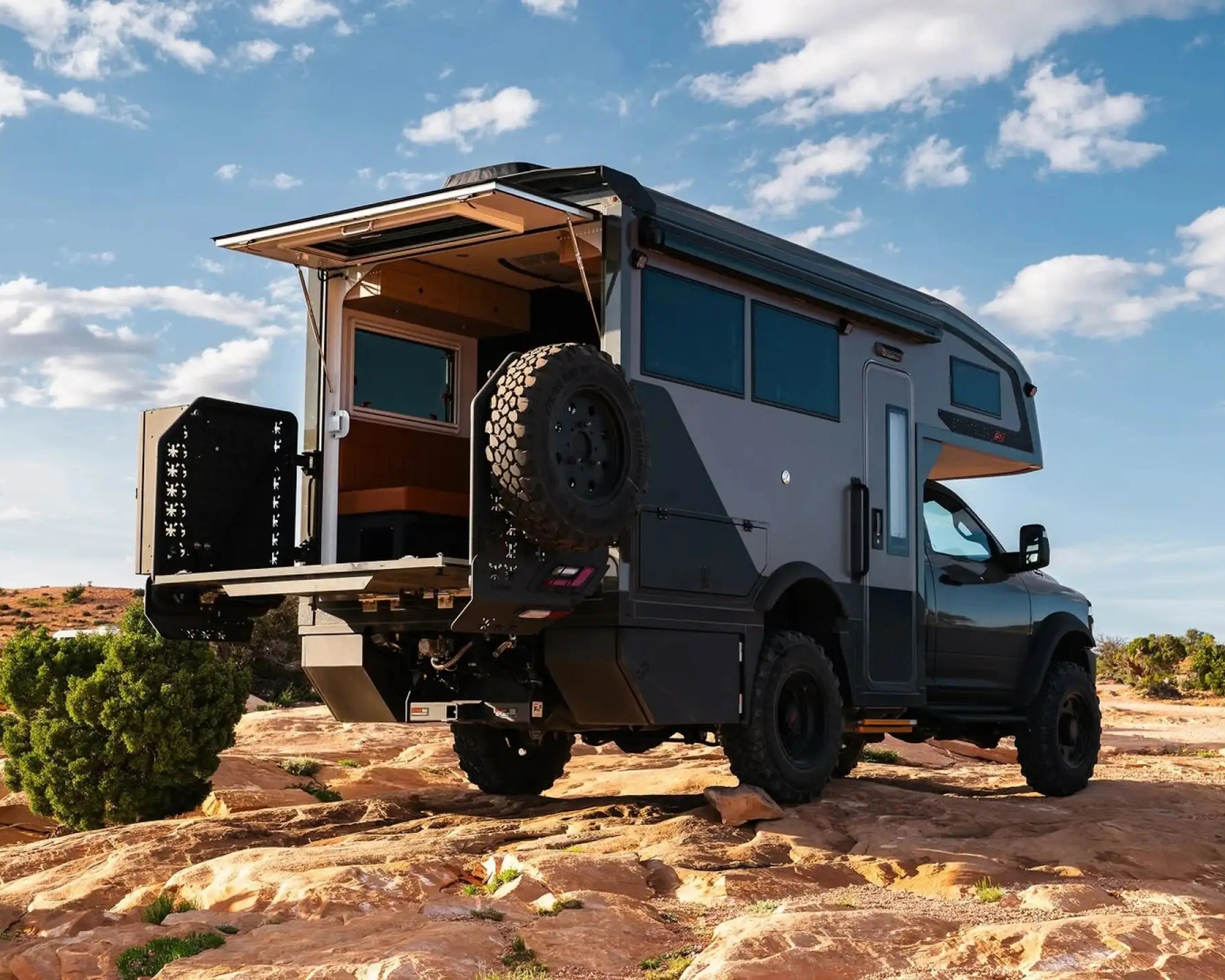 Off-road camper truck with rear storage compartment open, parked on rocky terrain under blue sky.