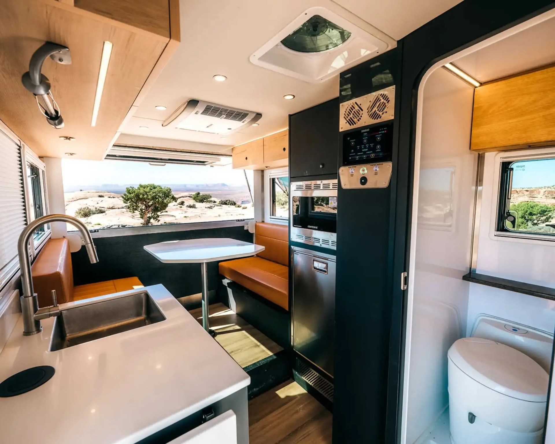 Compact RV interior with a kitchen sink, orange cushioned seating, a table, built-in appliances, and a small bathroom with a toilet.