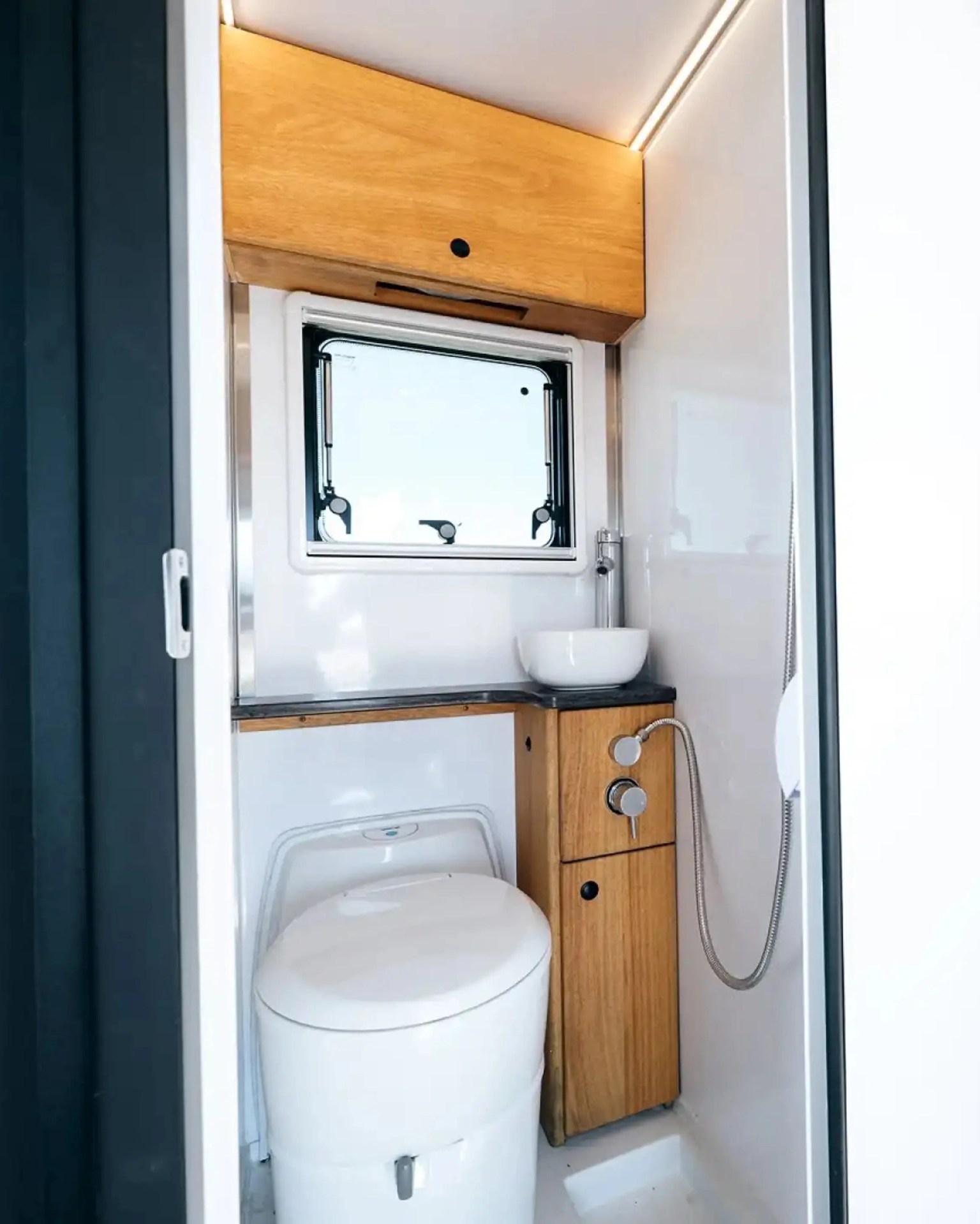 Compact white toilet with wooden cabinetry, black countertop, round white vessel sink, and small window in a bathroom.