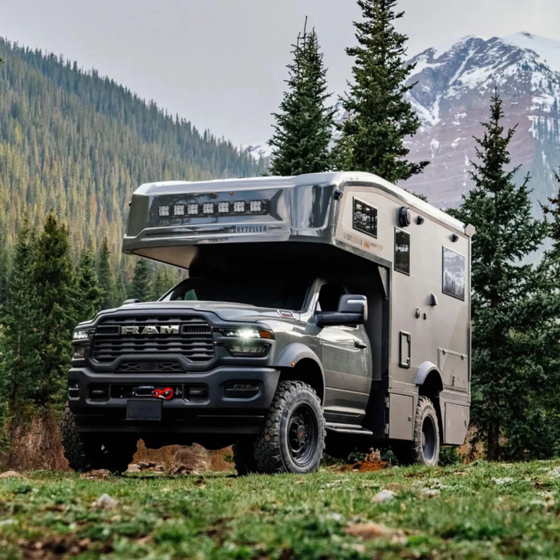 Gray RAM truck with a large camper mounted on the back, parked on grassy terrain with pine trees and snow-capped mountains in the background.