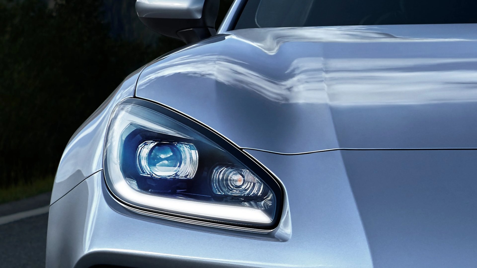 Close-up of a silver car's front left headlight and hood with reflections of clouds.
