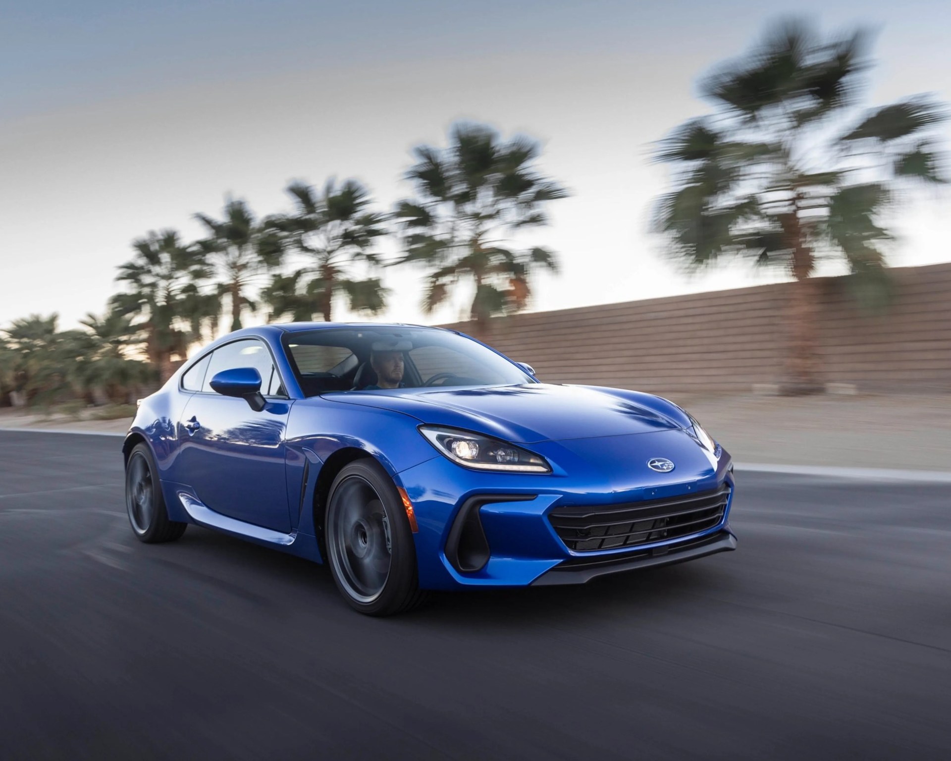 Blue Subaru sports car driving on a road with palm trees and a brick wall in the background.