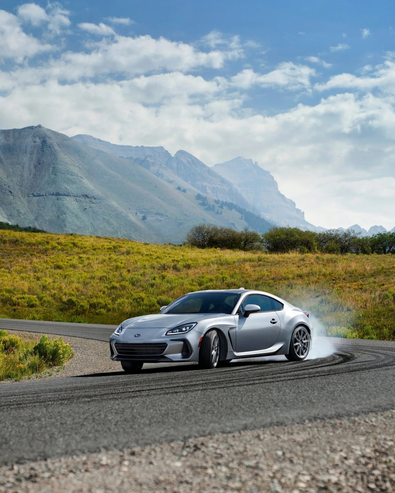 Silver Subaru sports car drifting on a mountain road with tire smoke and grassy hills in the background.