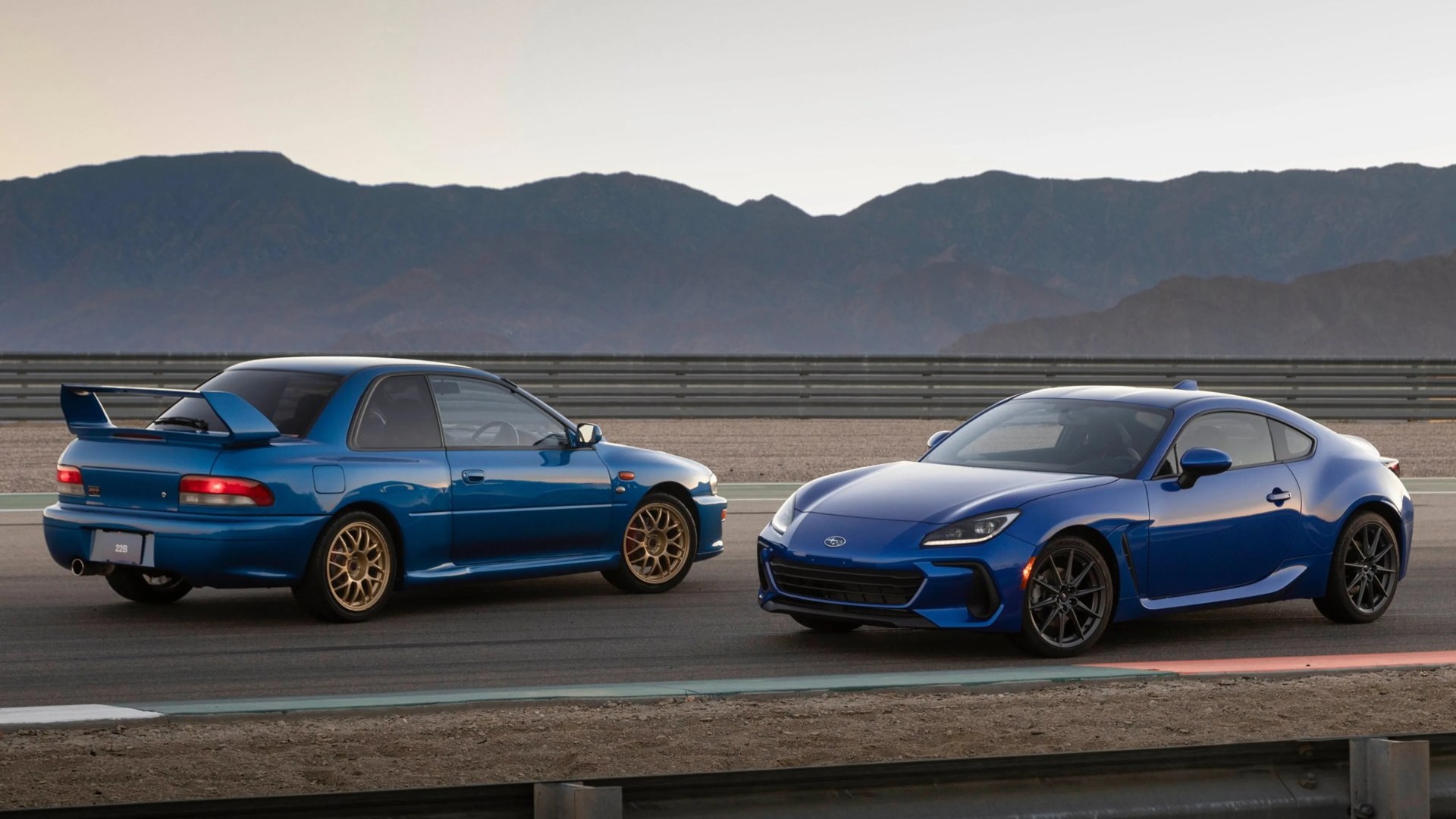 Two blue Subaru sports cars, one classic with gold wheels and a large rear spoiler, and one modern with black wheels, parked side by side on a racetrack with mountains in the background.