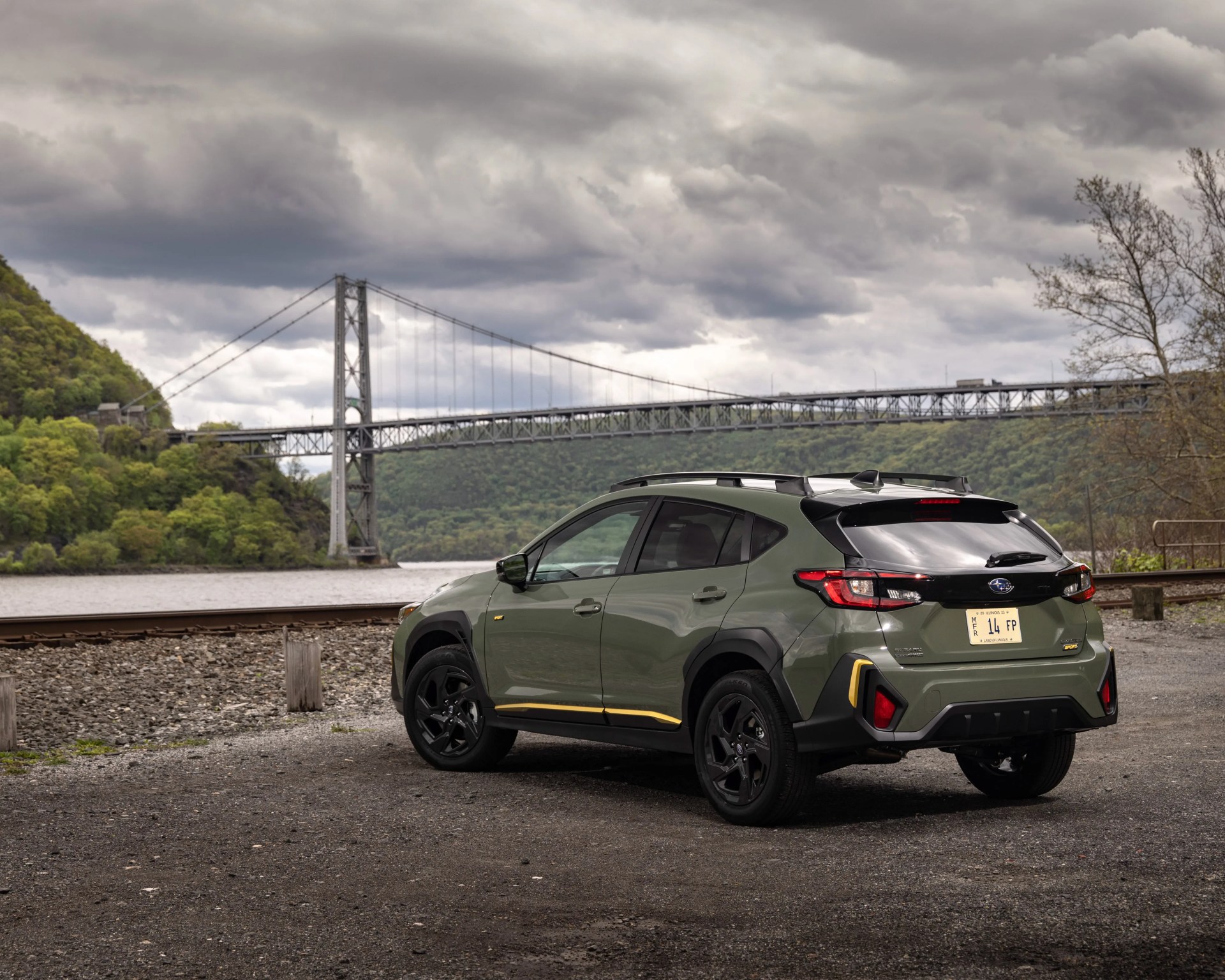 Green Subaru SUV with black and yellow trim parked near a river with a suspension bridge and cloudy sky in the background.