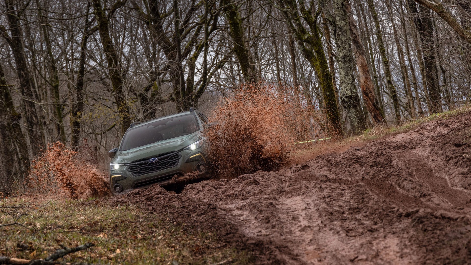 Green Subaru SUV driving through muddy forest trail splashing mud on both sides.