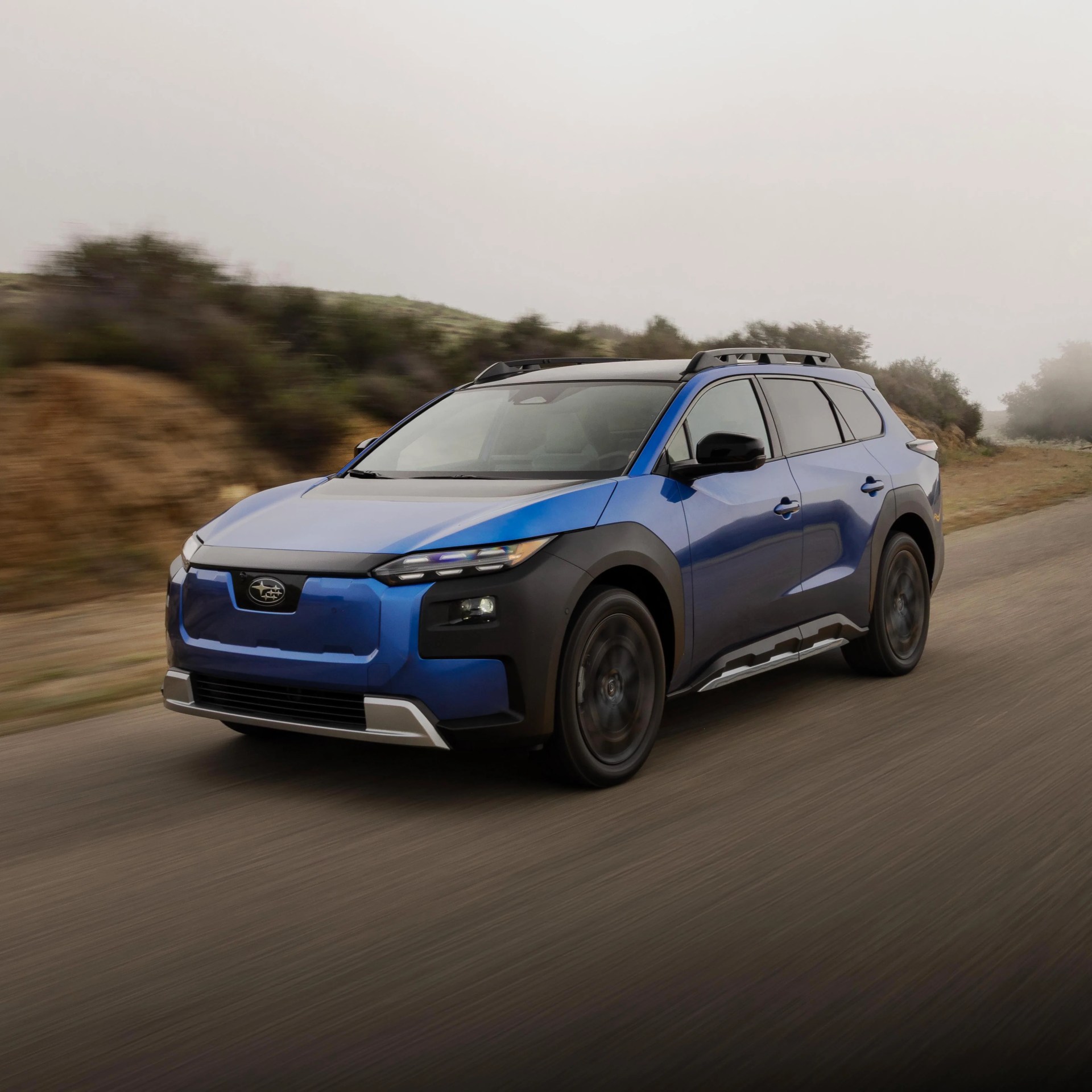 Blue Subaru SUV with black roof rails driving on a dirt road in a foggy, rural area.