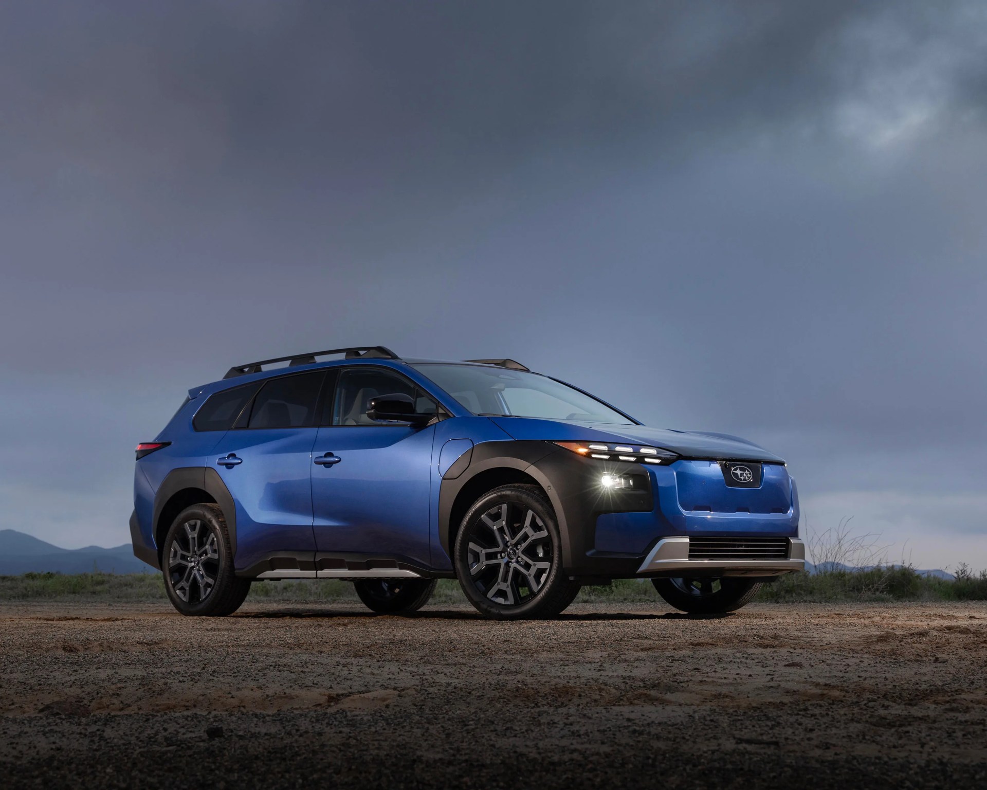 Blue Subaru SUV with black trim and roof rails parked on dirt under cloudy sky.