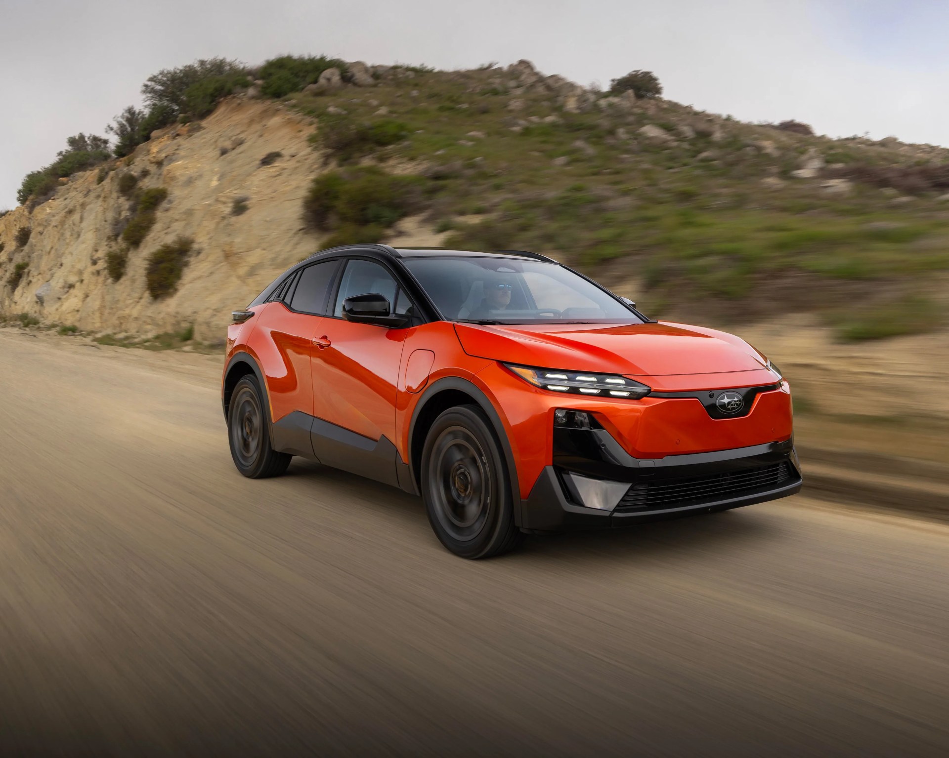 Orange Subaru SUV with black roof driving on a dirt road near a rocky hillside.