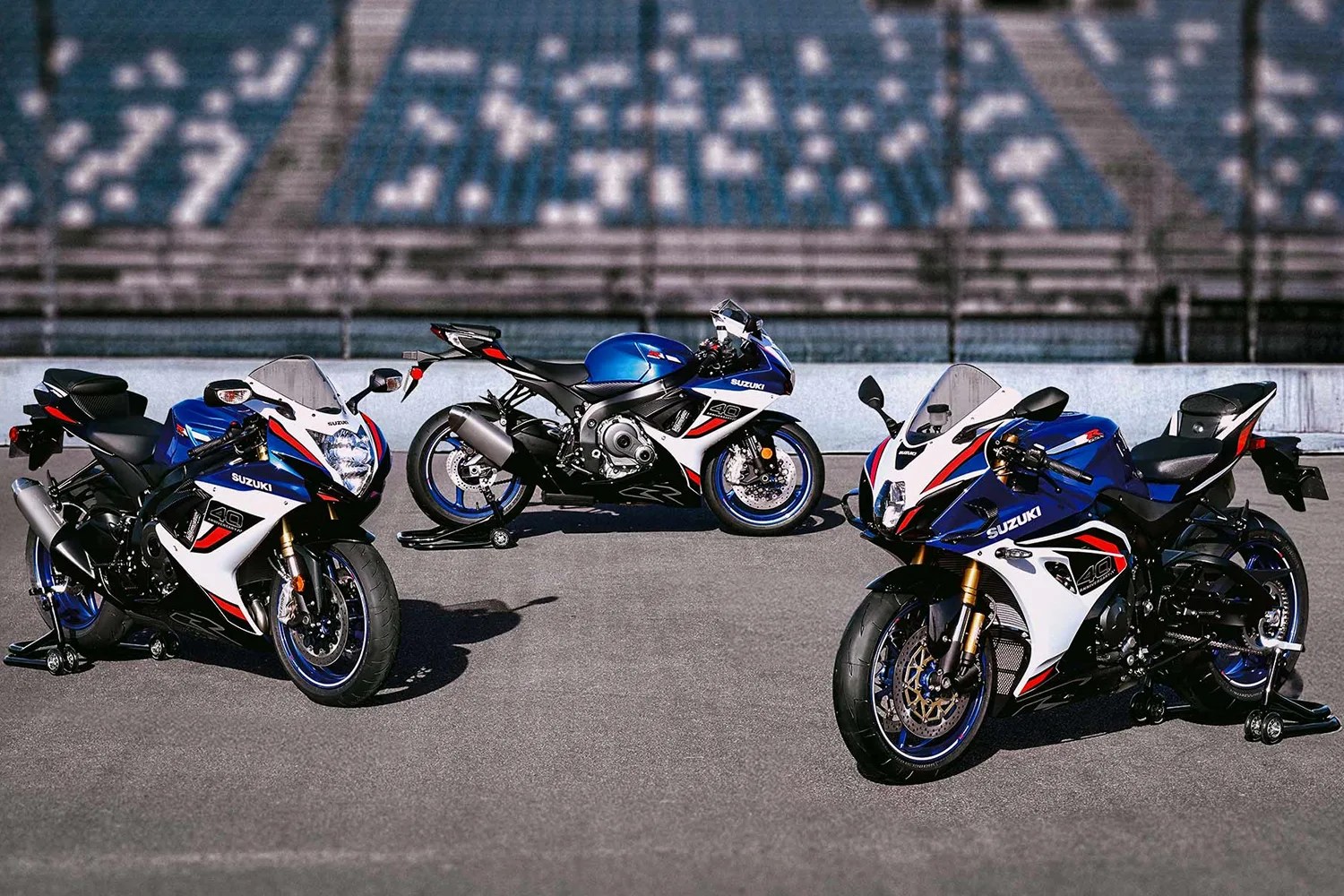 Three blue, white, and black Suzuki sport motorcycles with "40" decals parked on a racetrack with empty bleachers in the background.