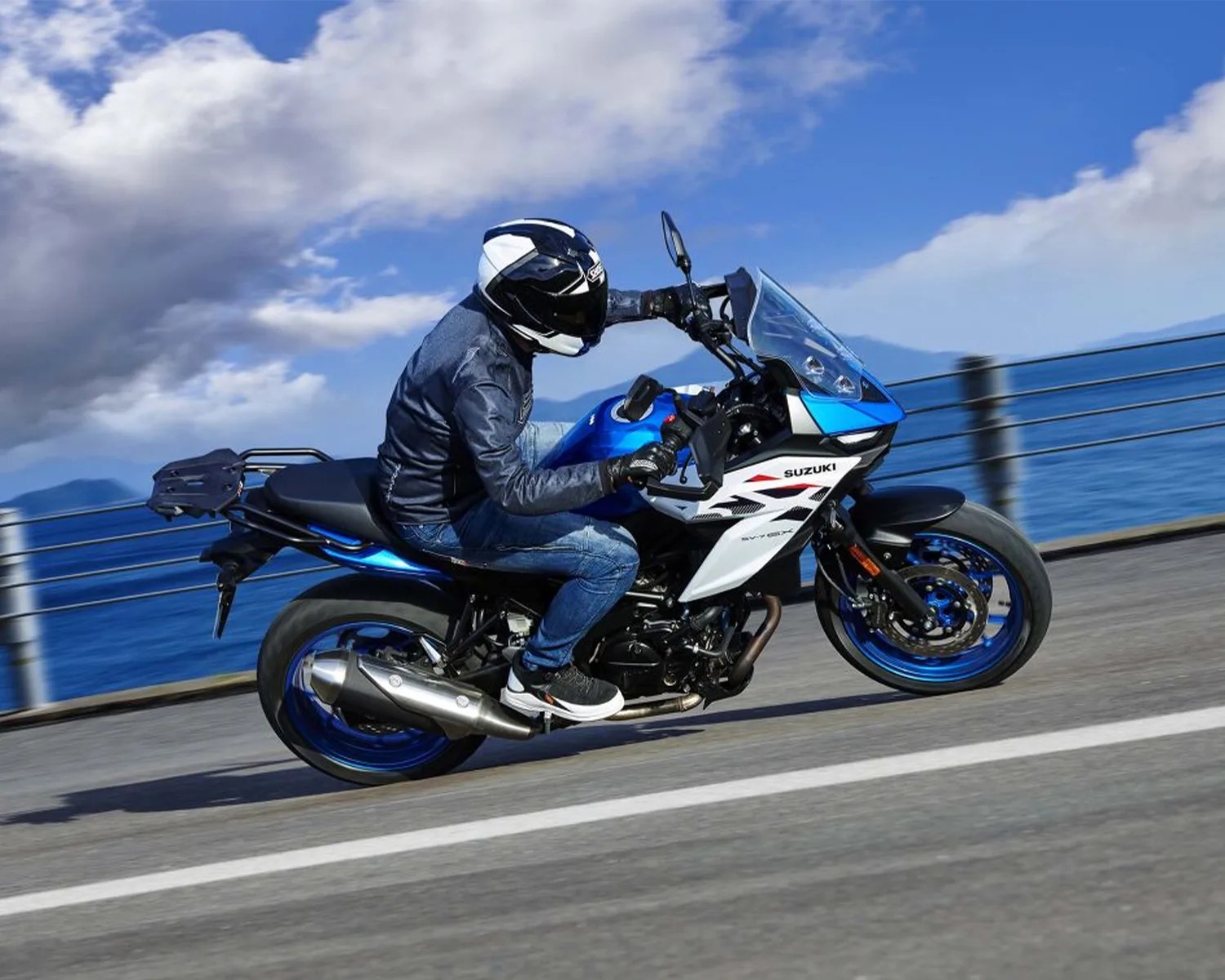 Person wearing a black and white helmet riding a blue and white Suzuki motorcycle on a road by the water.