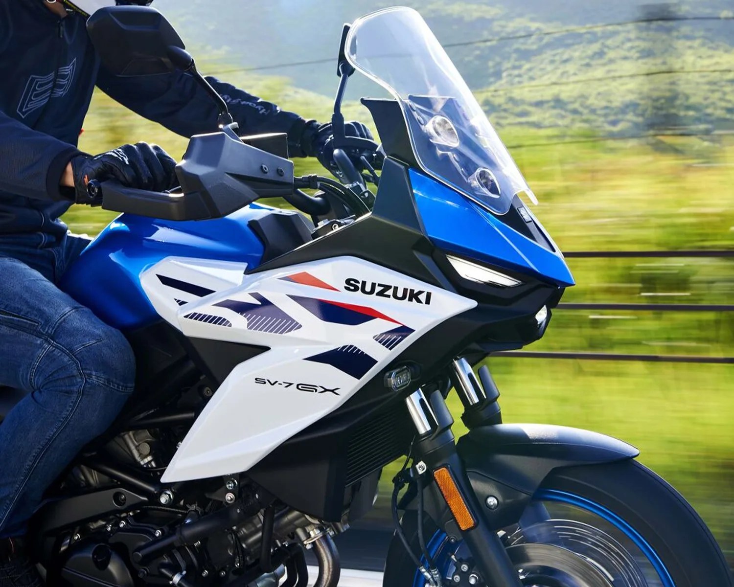 Blue and white Suzuki SV-7 GX motorcycle front with clear windshield and rider in gloves and jeans.