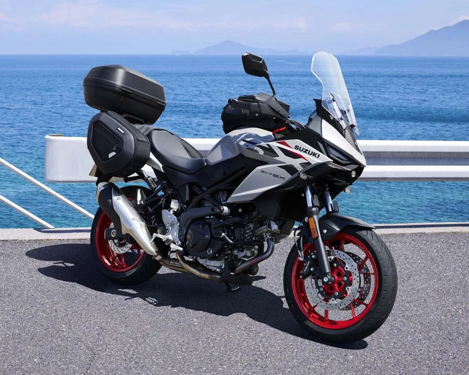 White and black Suzuki V-Strom 650XT motorcycle with red wheels and luggage boxes parked by the sea.