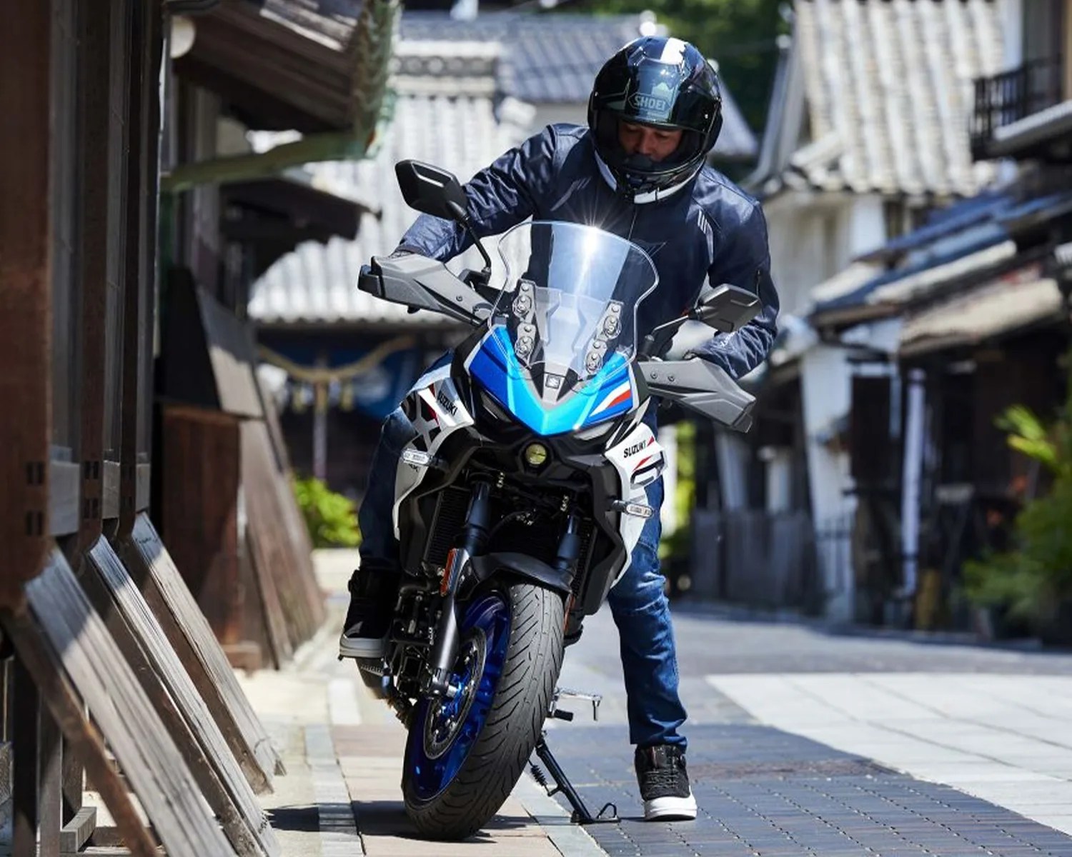 Person wearing a black helmet and jacket sitting on a blue and white Suzuki motorcycle on a paved street.