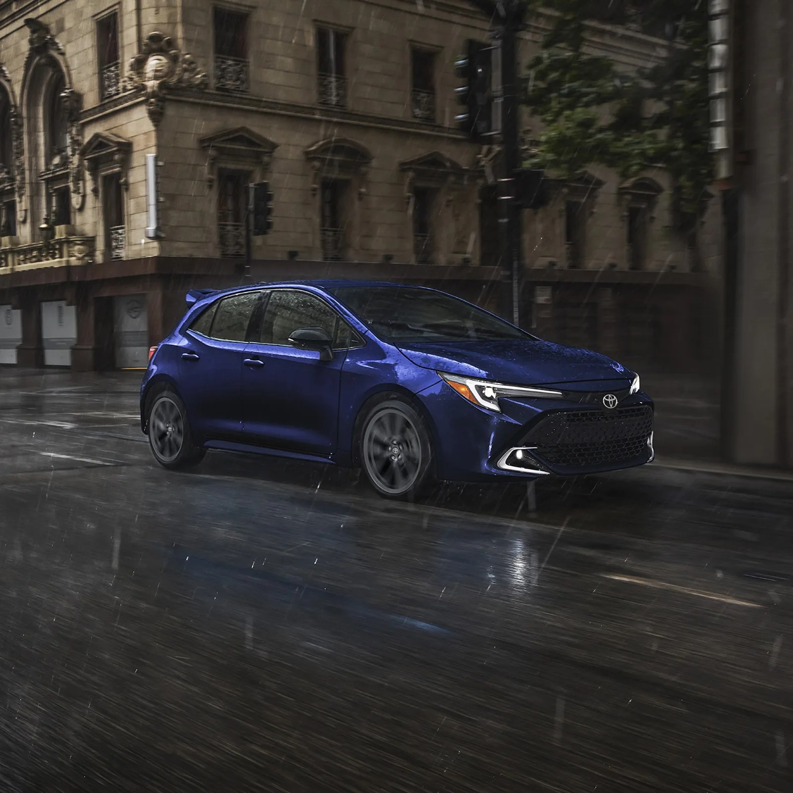 Blue Toyota hatchback driving on a wet city street in the rain.