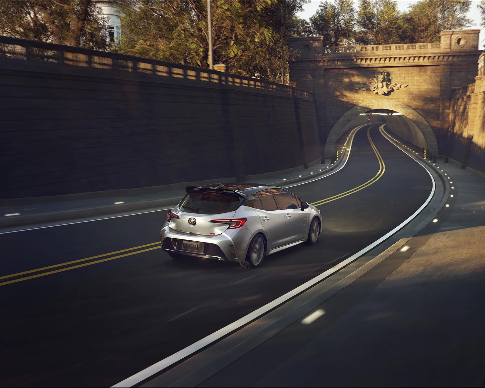 Silver Toyota hatchback with black roof driving on a curved road entering a stone tunnel.